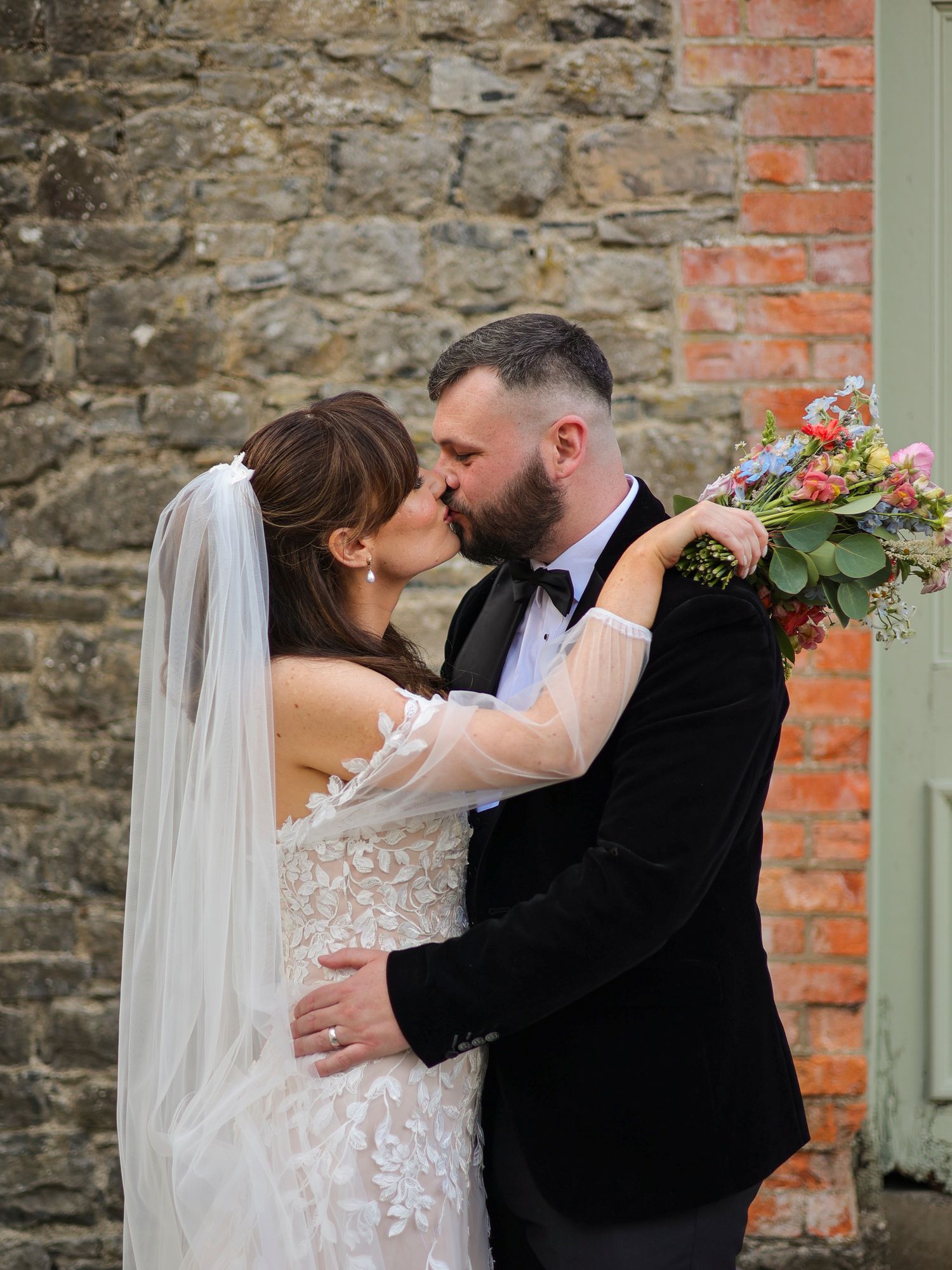 Bride and groom sharing a romantic kiss while holding a colorful bouquet against a rustic brick wall.