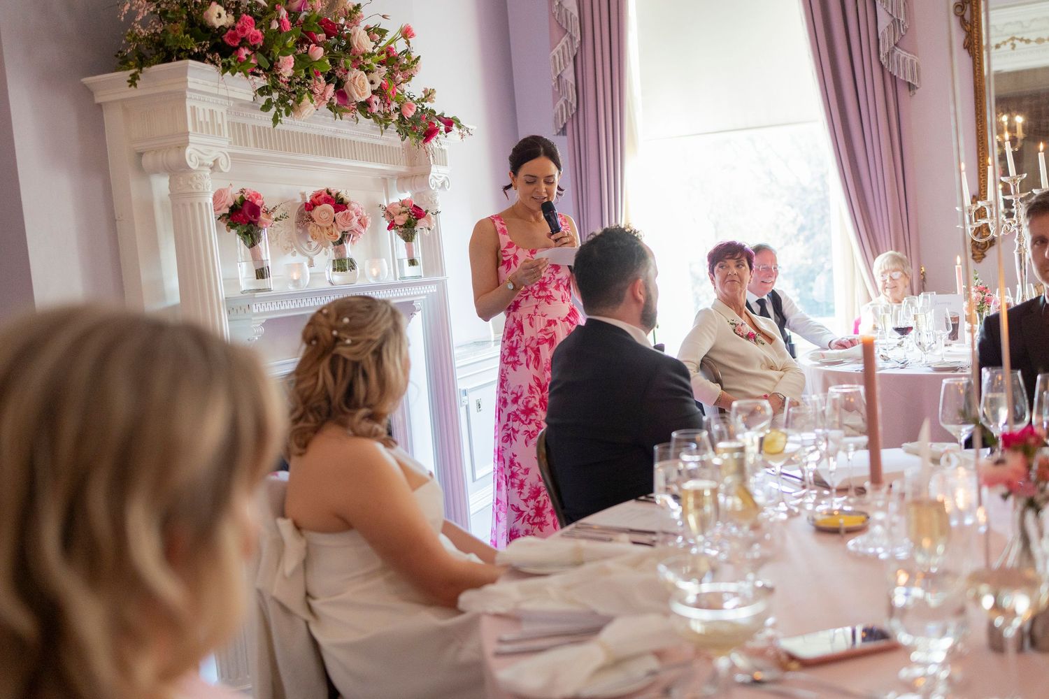 Woman in floral dress giving a speech at an elegant wedding reception dinner with guests seated at decorated tables.