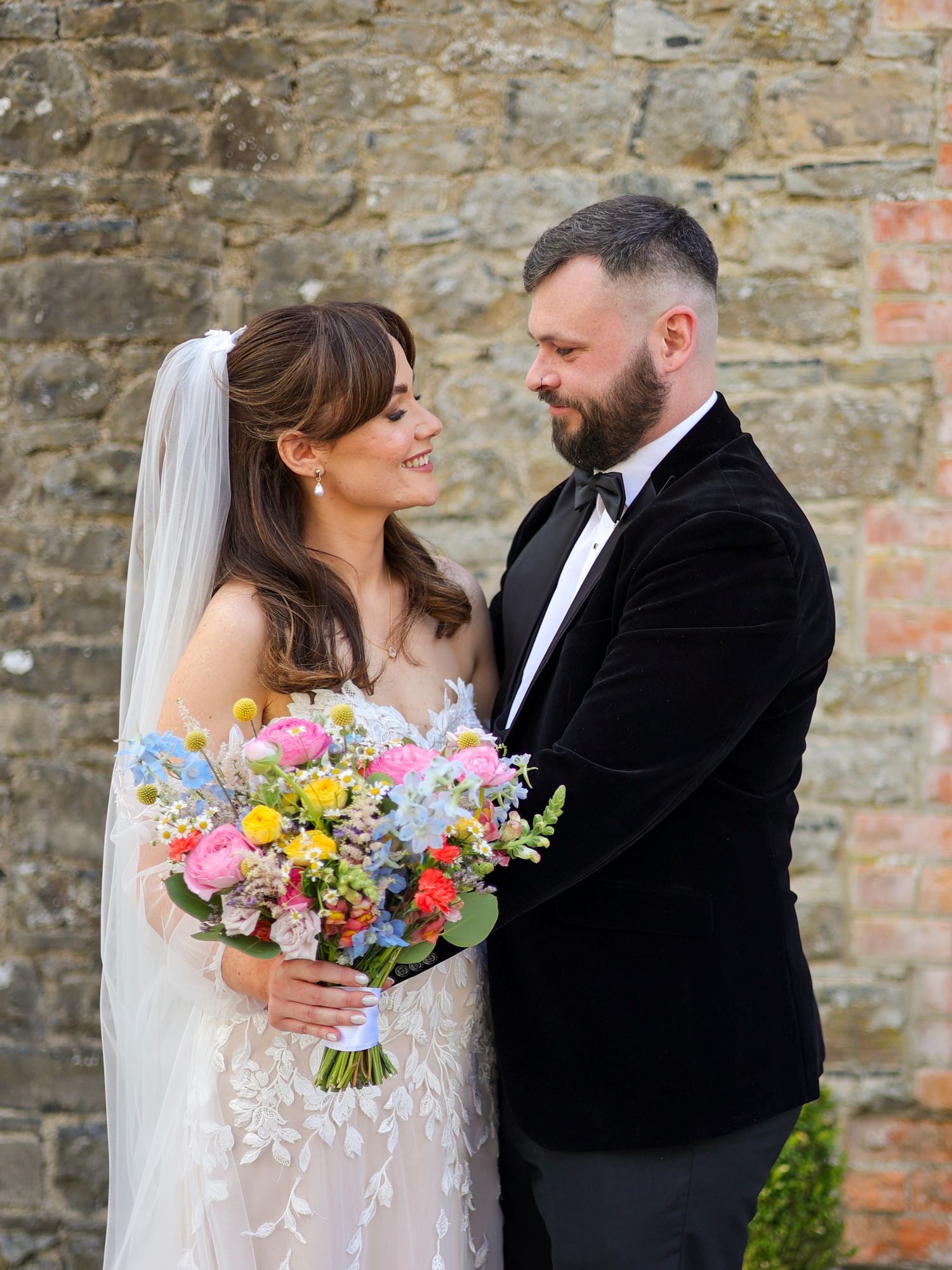 Bride and groom share a romantic gaze beside a stone wall, bride holding a vibrant wildflower bouquet.