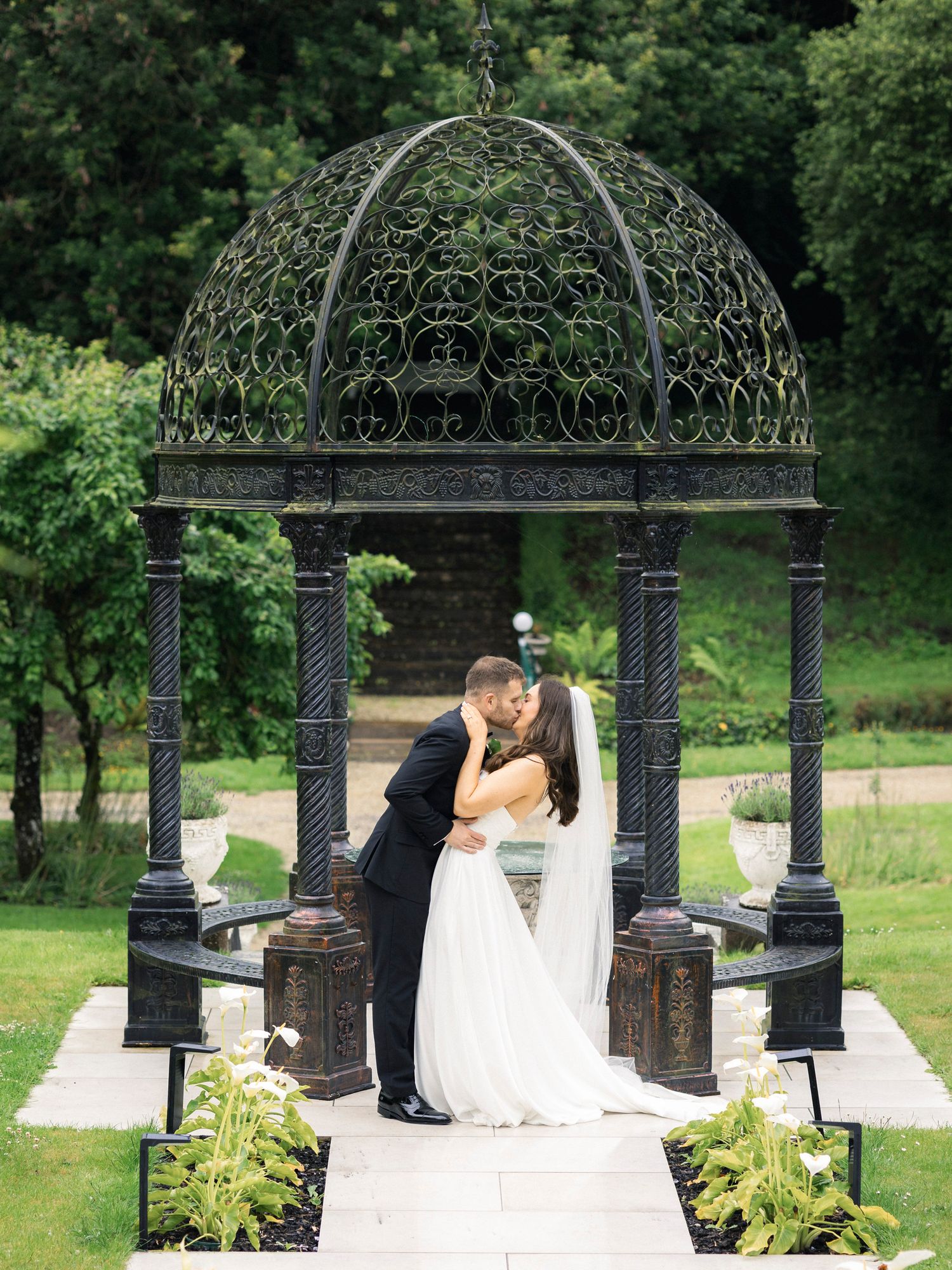 Bride and groom share a romantic kiss under an ornate black iron gazebo surrounded by lush green gardens.