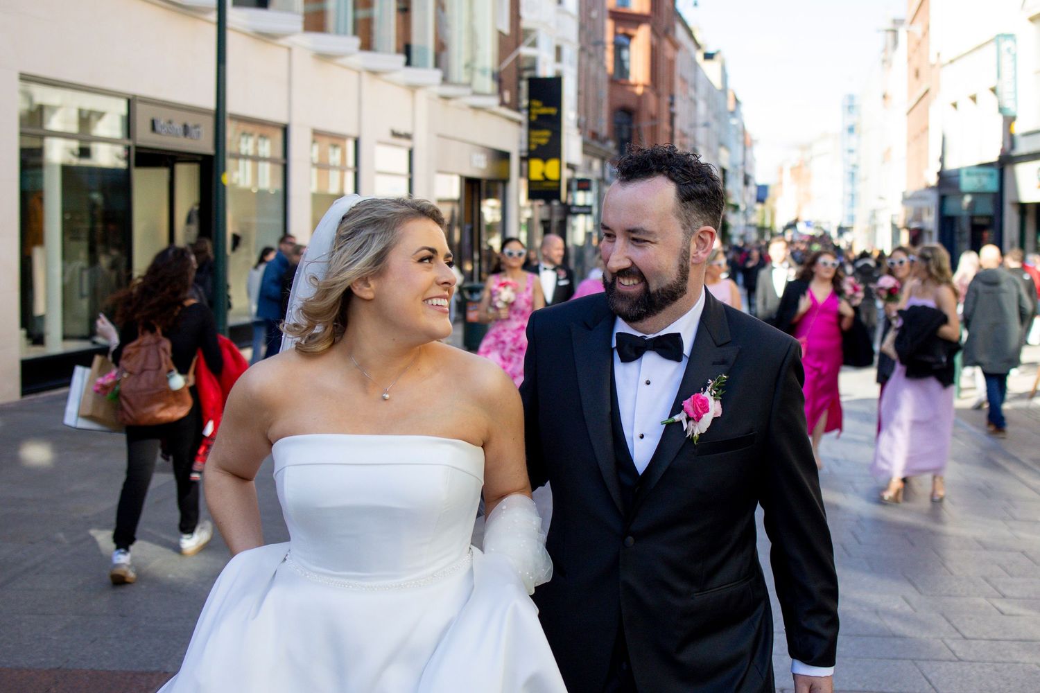 Bride and groom walking hand in hand through a busy city street, smiling at each other on their wedding day.