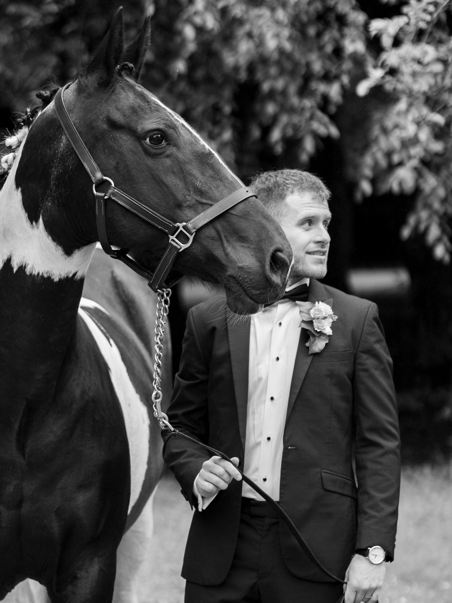 Black and white photo of a groom in a tuxedo standing beside a paint horse outdoors.