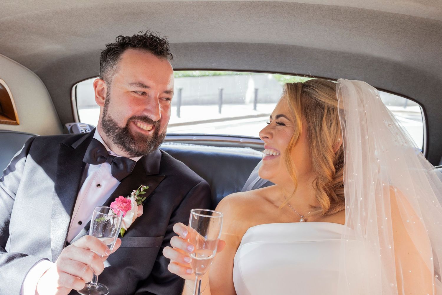 Newlyweds toast with champagne flutes, smiling joyfully together in the back seat of a classic car.