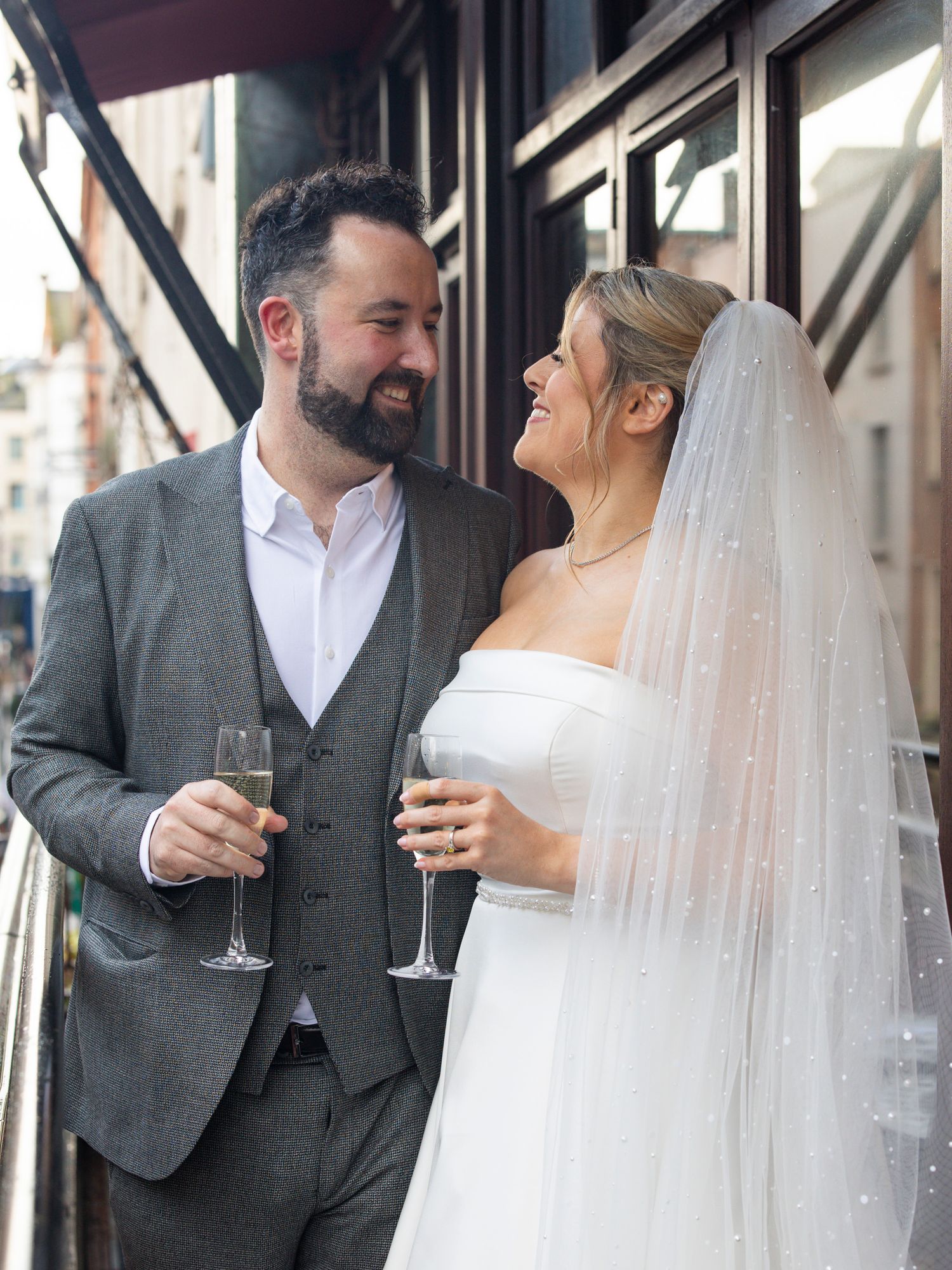 Newlywed couple smiling at each other holding champagne glasses, bride in white gown with veil, groom in grey suit.
