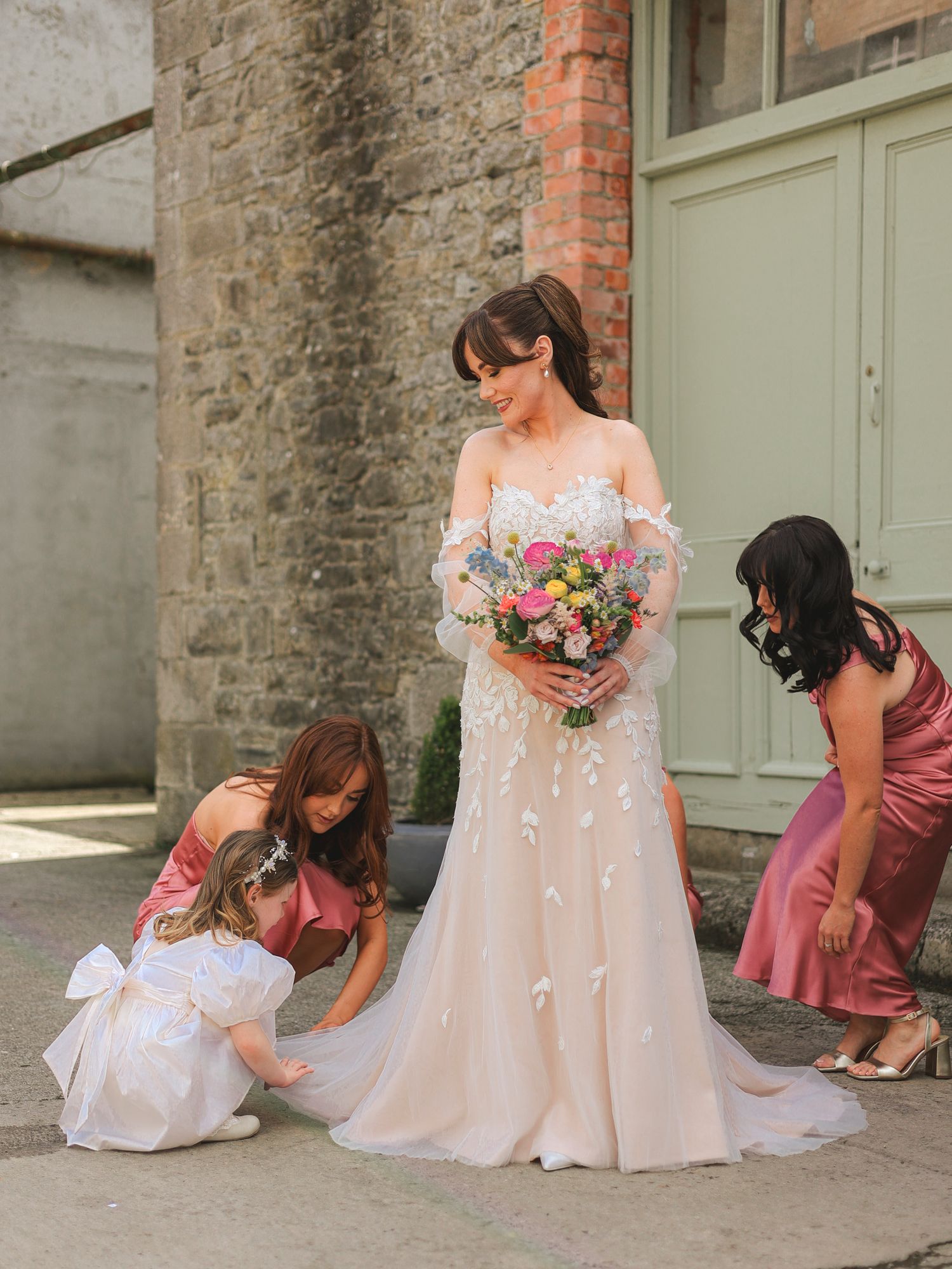 Bride in blush off-shoulder wedding gown holds colorful bouquet while flower girls in pink and white dresses adjust her train.