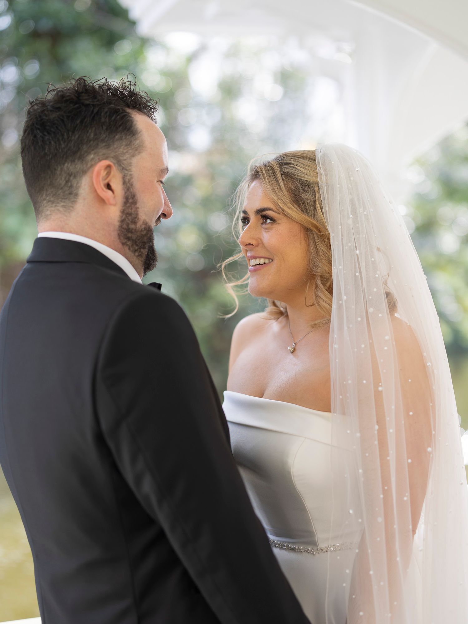Bride and groom smiling at each other during outdoor wedding ceremony, bride wearing white dress and veil.