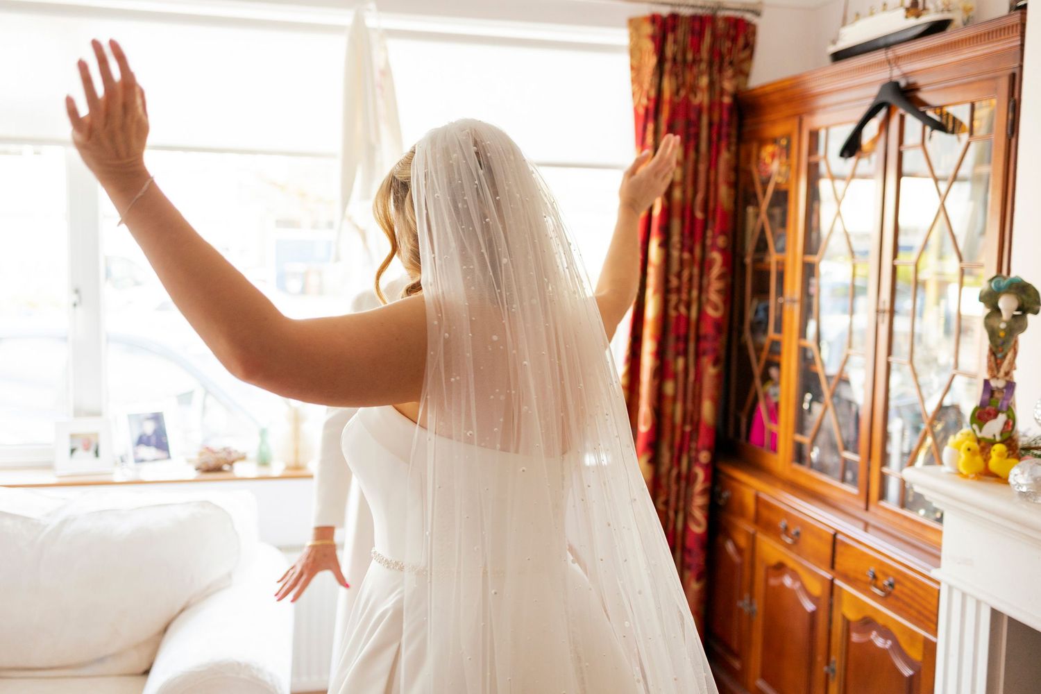 Bride in white wedding gown and veil stands with arms raised by a bright window in a elegant room.