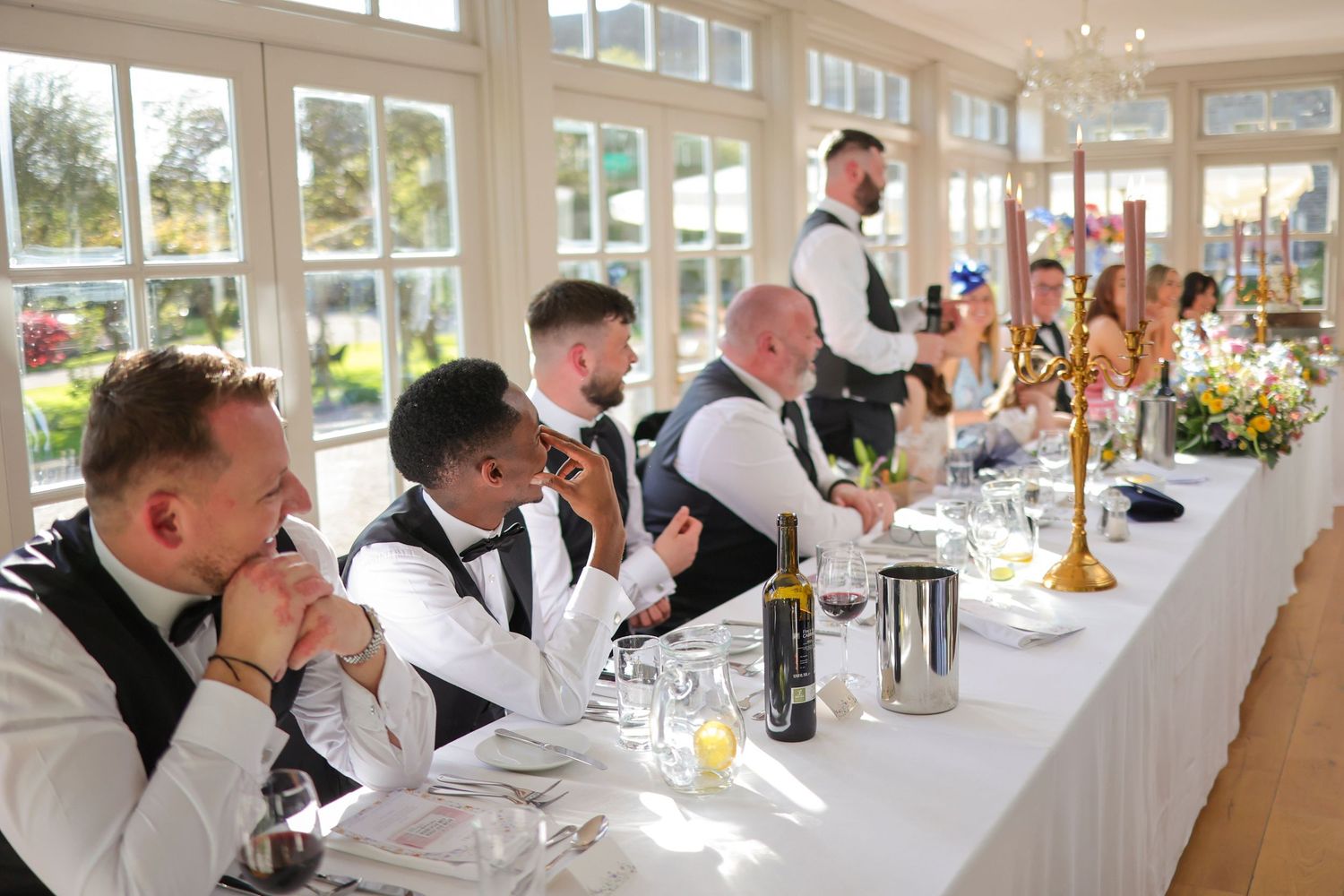 Groomsmen in black waistcoats seated at a wedding reception head table in a bright conservatory dining room.