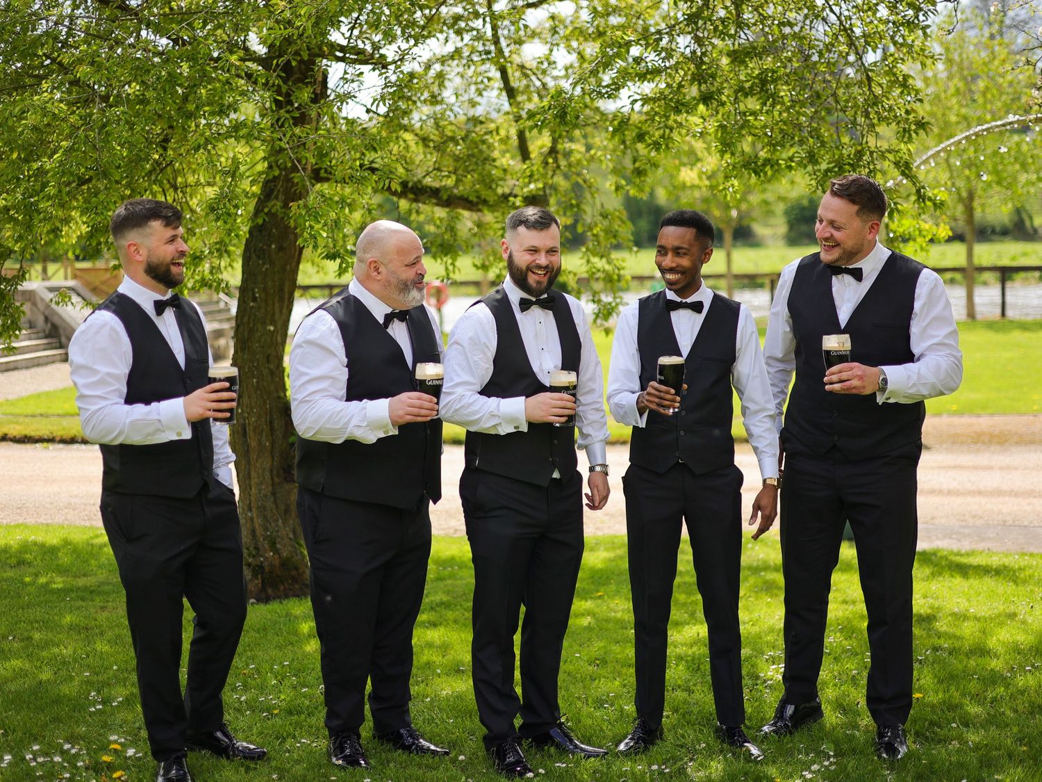 Five groomsmen in black tuxedo vests and bow ties socializing outdoors under green trees at a wedding.