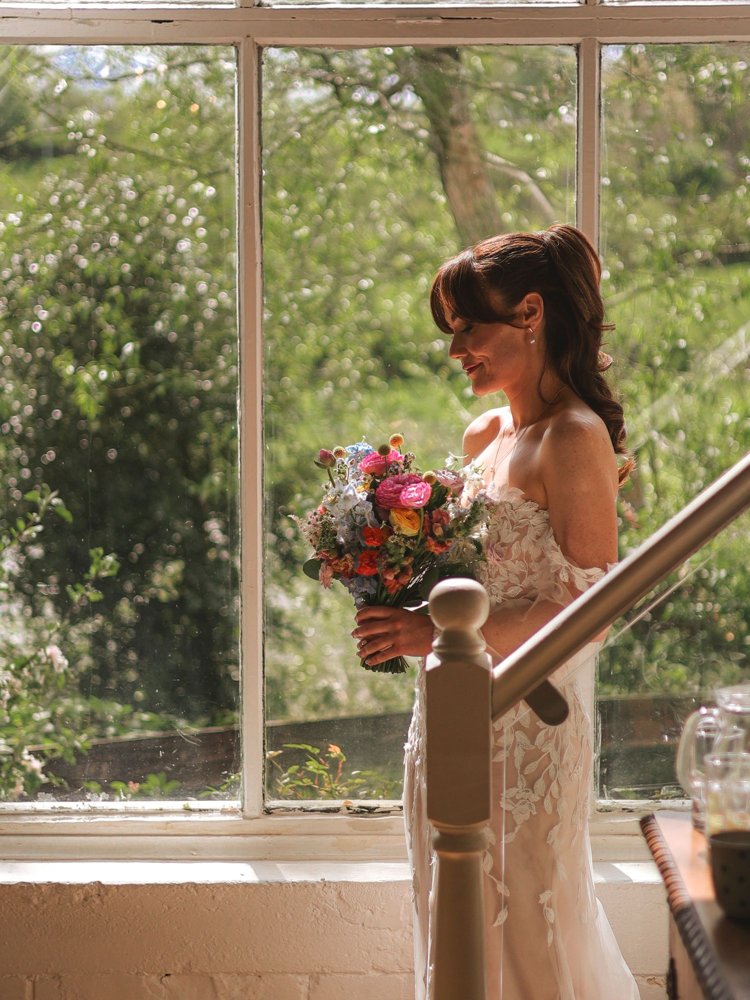Bride in lace gown holds colorful bouquet by sunlit window, smiling softly on wedding day.