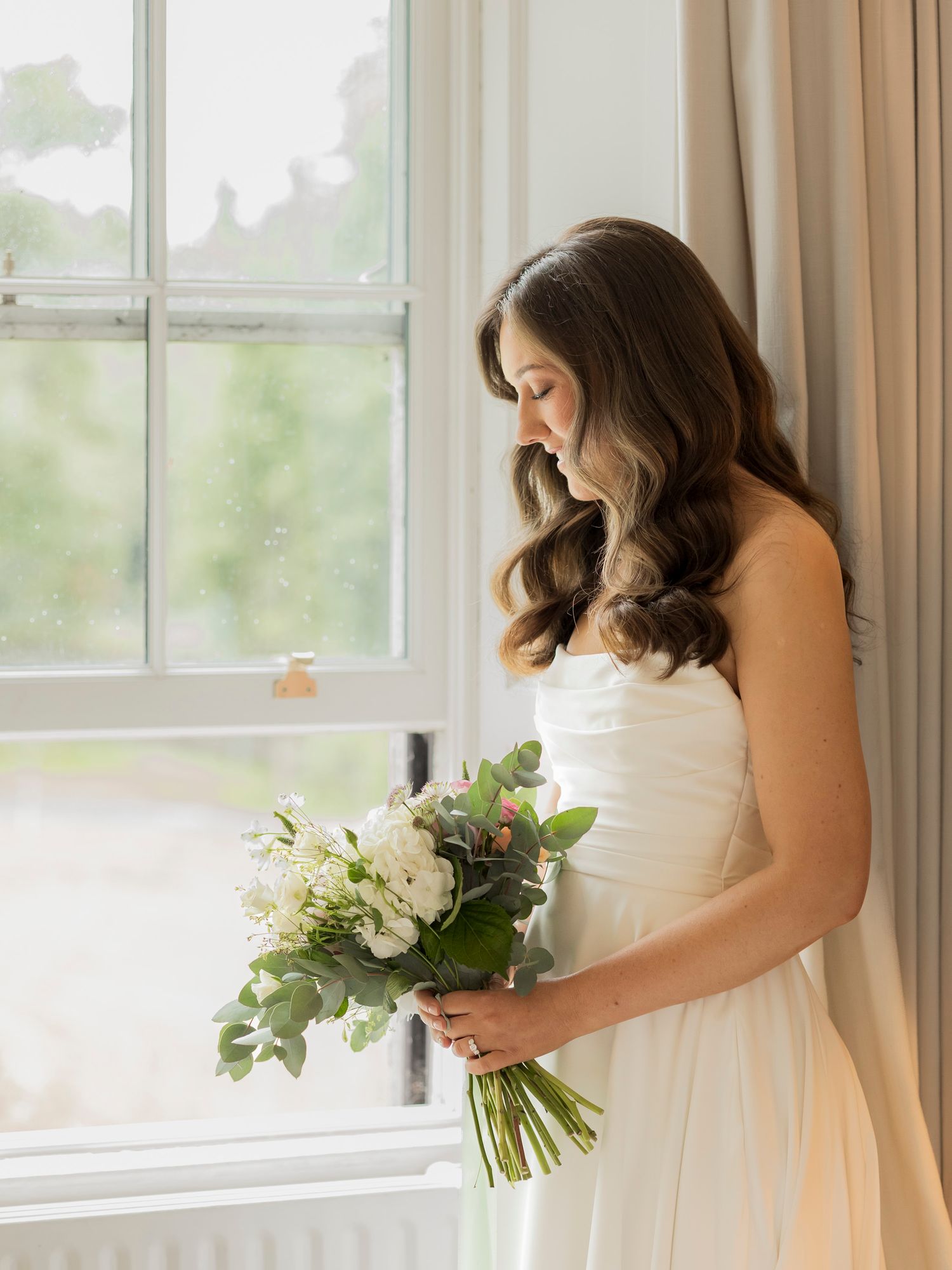 Bride in strapless white gown holding elegant white floral bouquet, standing by bright window with soft natural light.