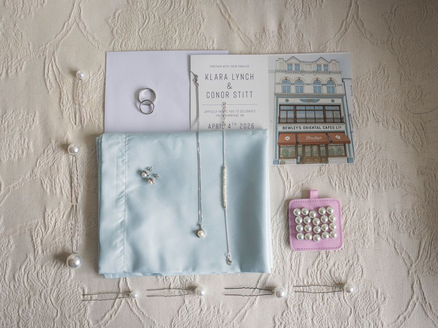 Flat lay of wedding stationery, pearl jewelry, light blue satin pouch, and hair accessories.