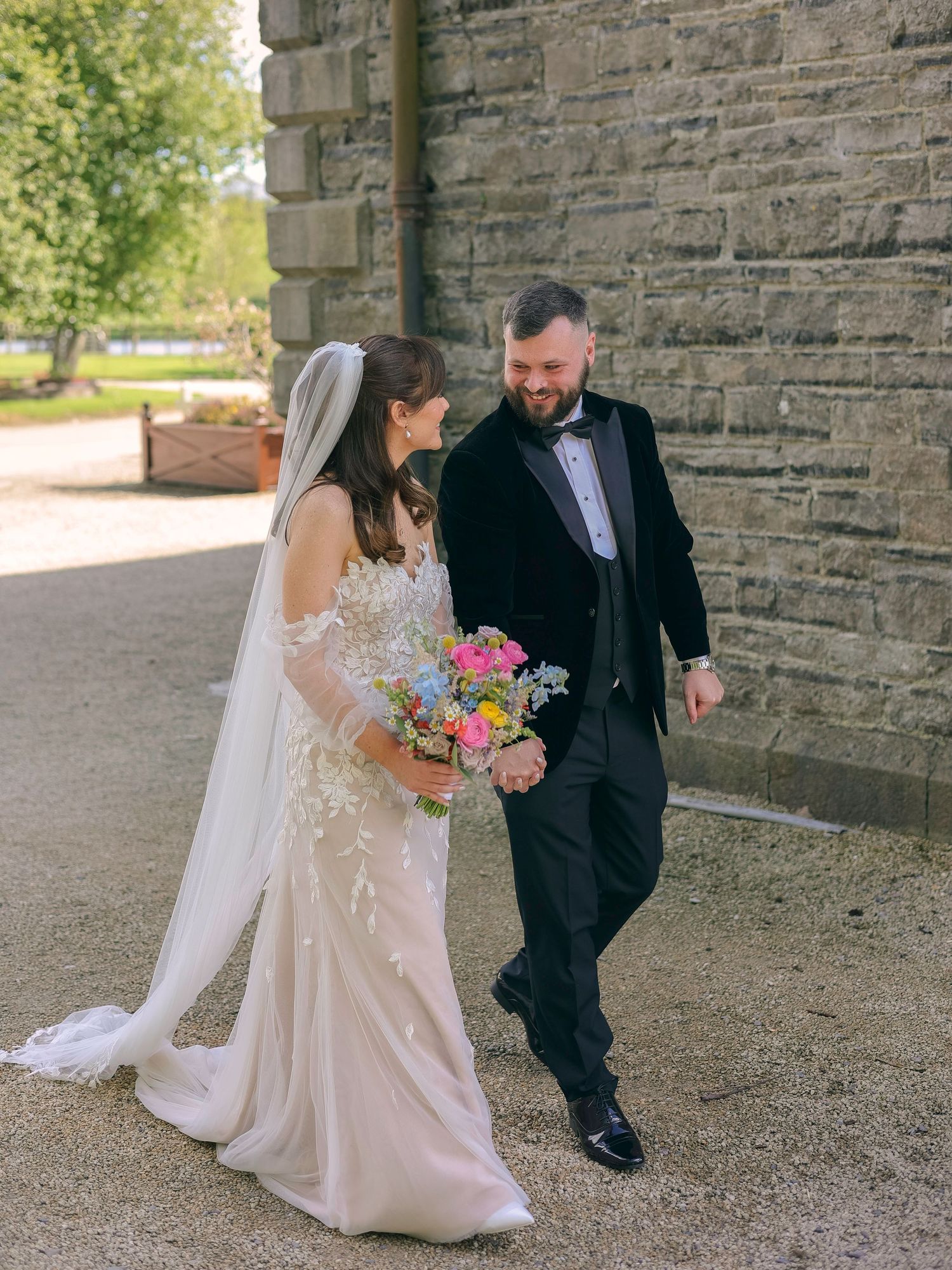 Bride in lace gown and groom in black velvet tuxedo walk and laugh beside a stone wall.
