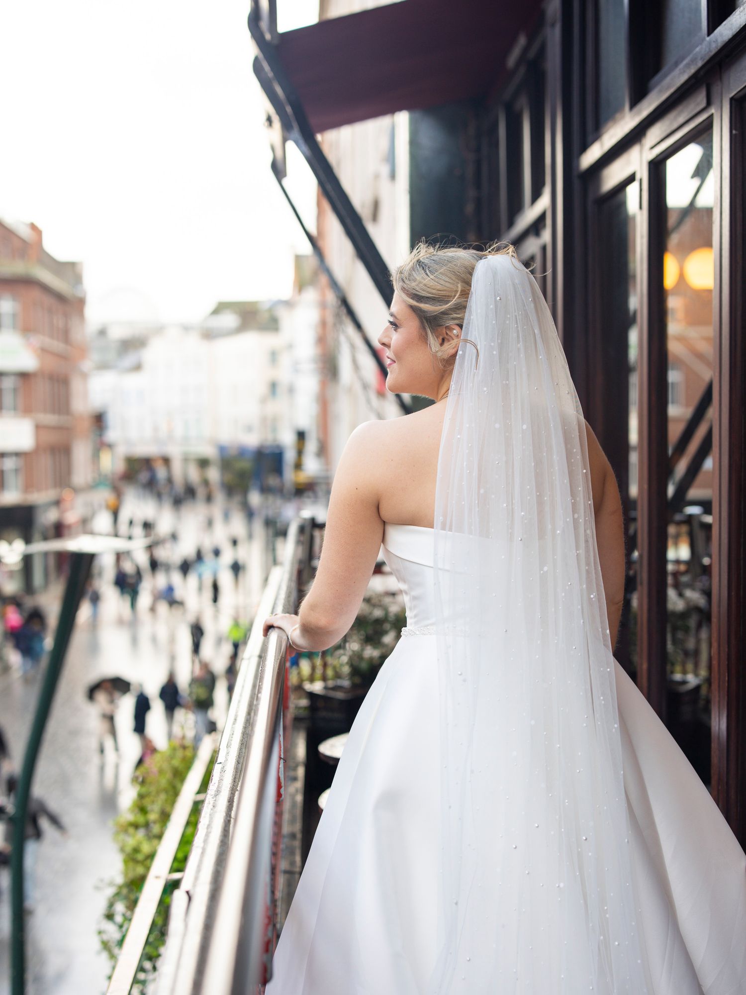 Bride in white strapless gown and veil stands on city balcony overlooking busy rainy street below.