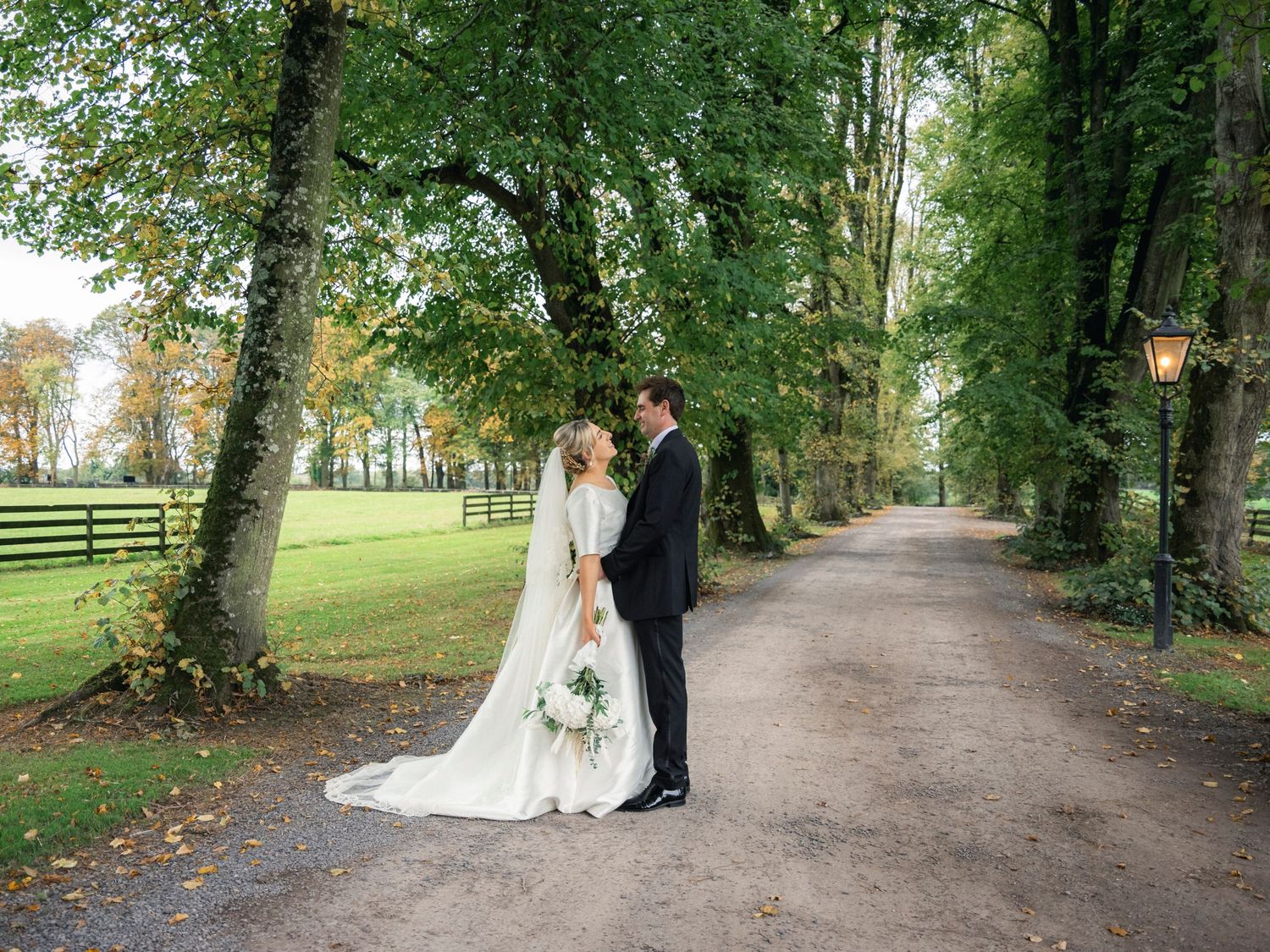 Bride and groom share tender moment on tree-lined country estate driveway in autumn.