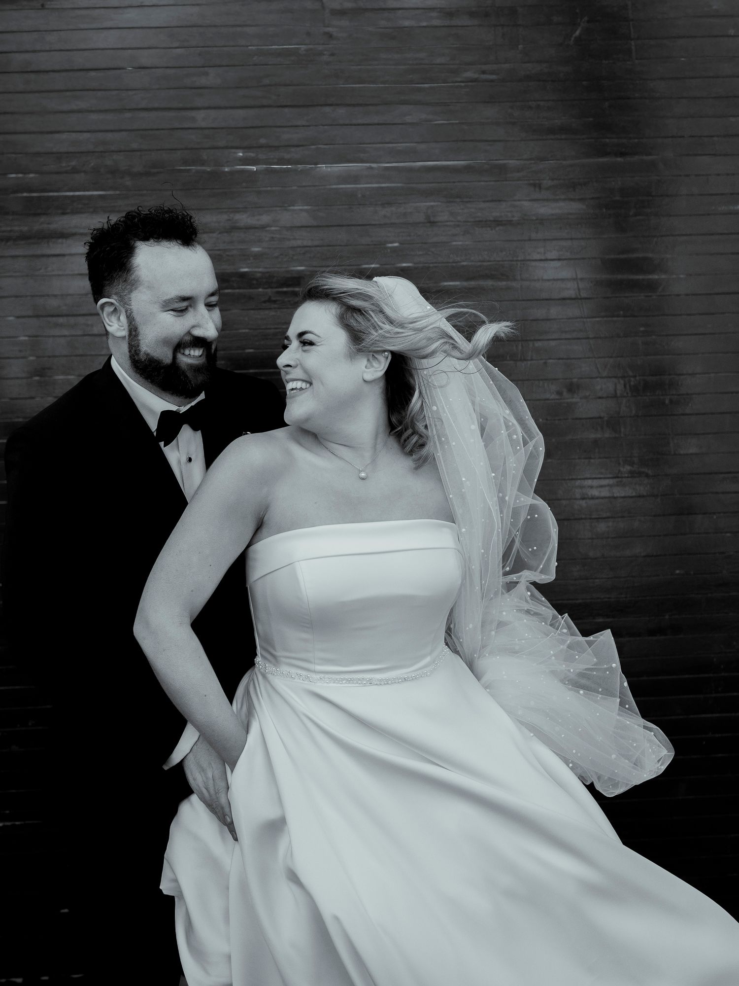 Black and white portrait of bride and groom with flowing veil against dark wooden wall.