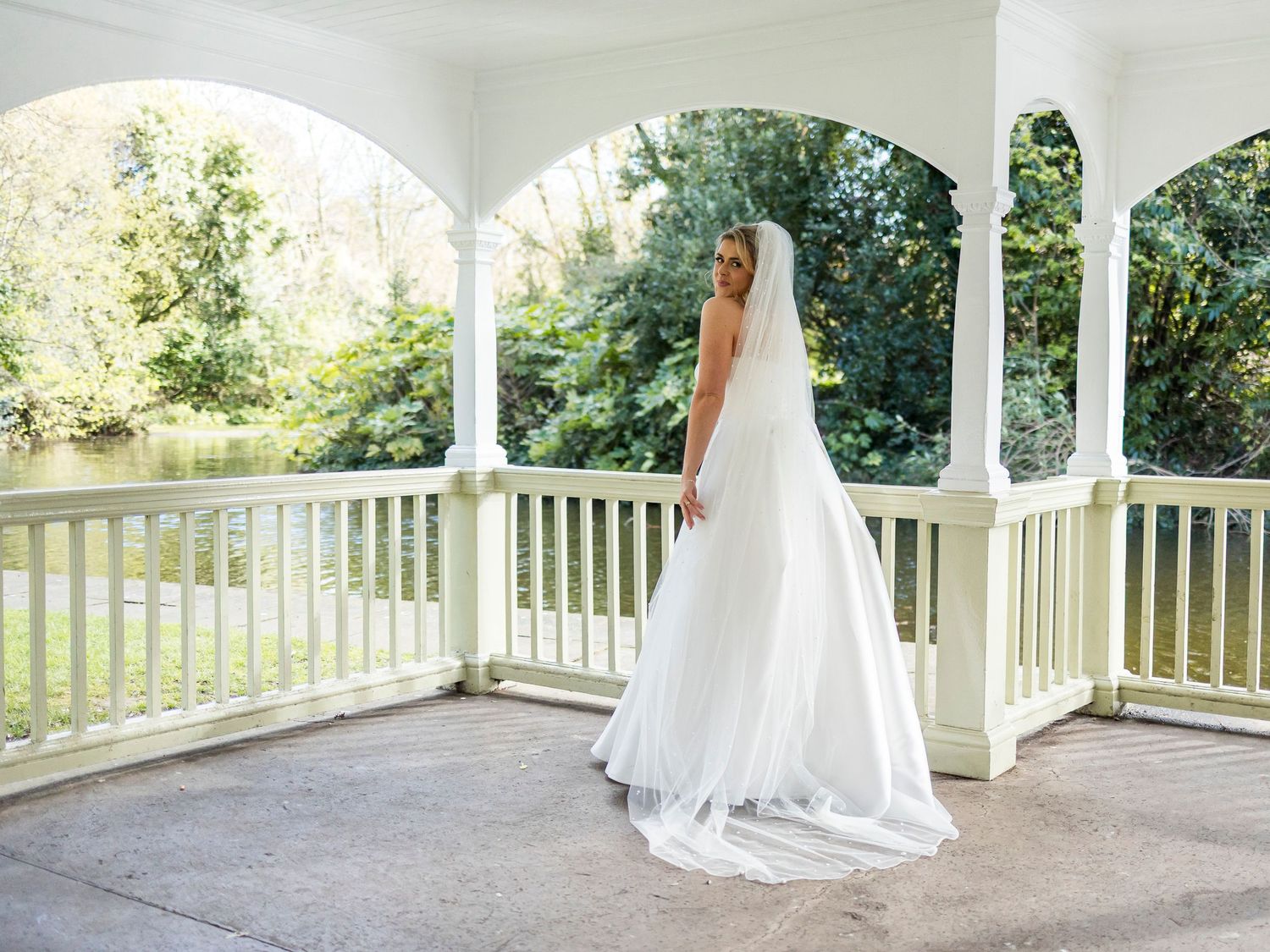 Bride in elegant white gown stands on covered porch overlooking green lawn.