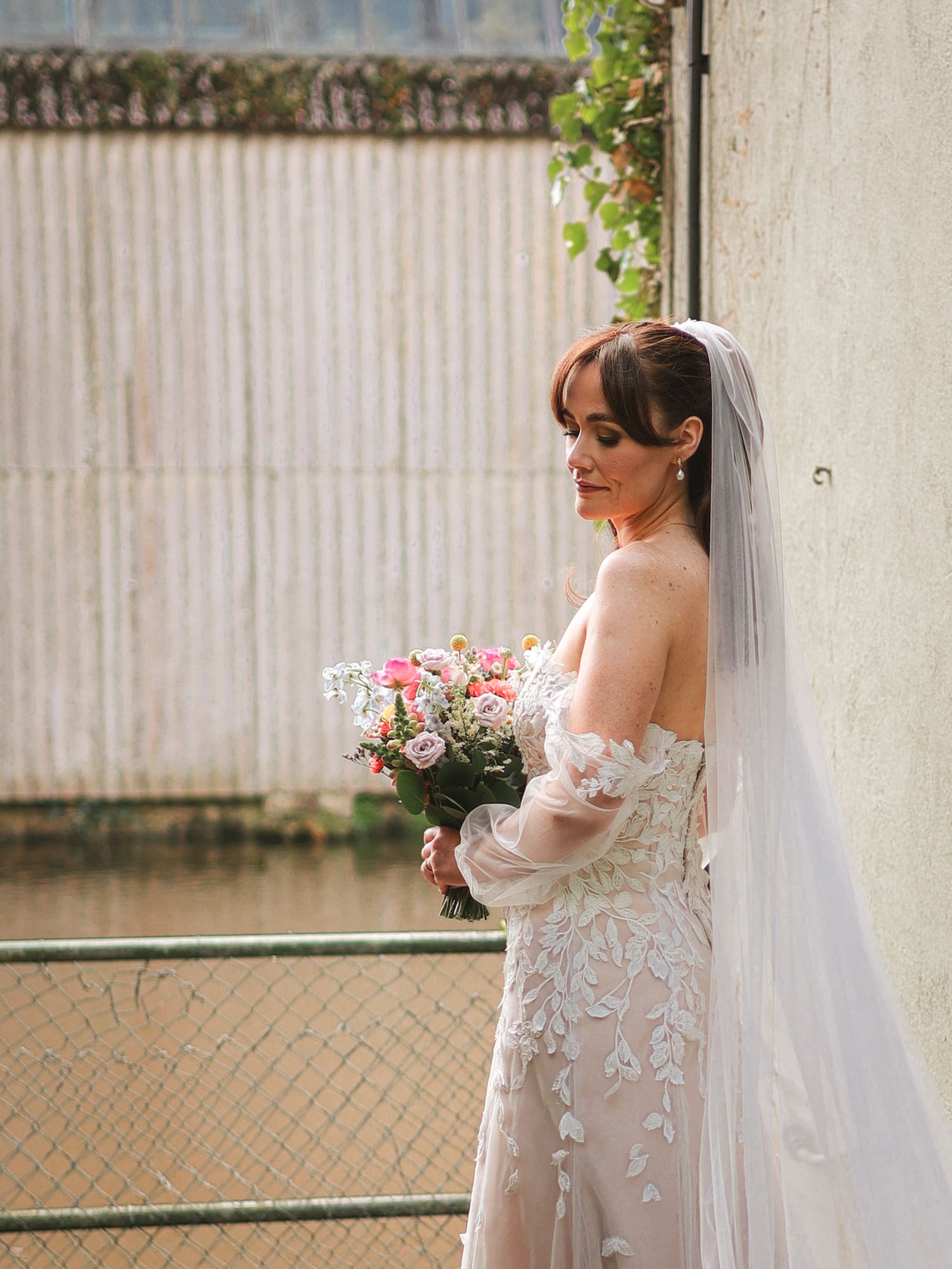 Elegant bride in lace off-shoulder gown and veil holds pink floral bouquet, posing outdoors in soft natural light.