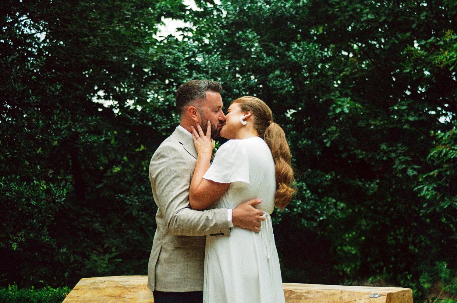 Newlywed couple sharing romantic kiss outdoors surrounded by lush green trees after wedding ceremony.