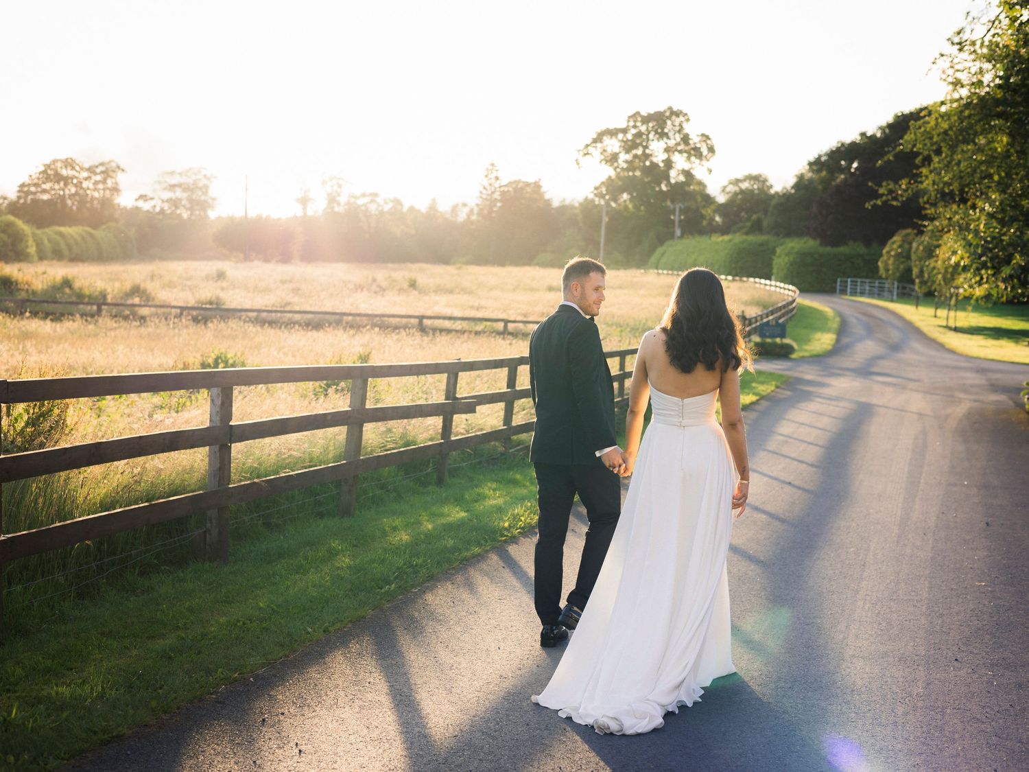 Bride and groom walking hand in hand down a sunlit country road beside a wooden fence at golden hour.