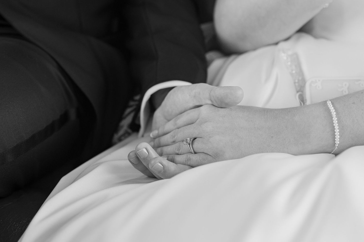 Black and white photo of a bride and groom holding hands, showing wedding rings and bridal gown detail.