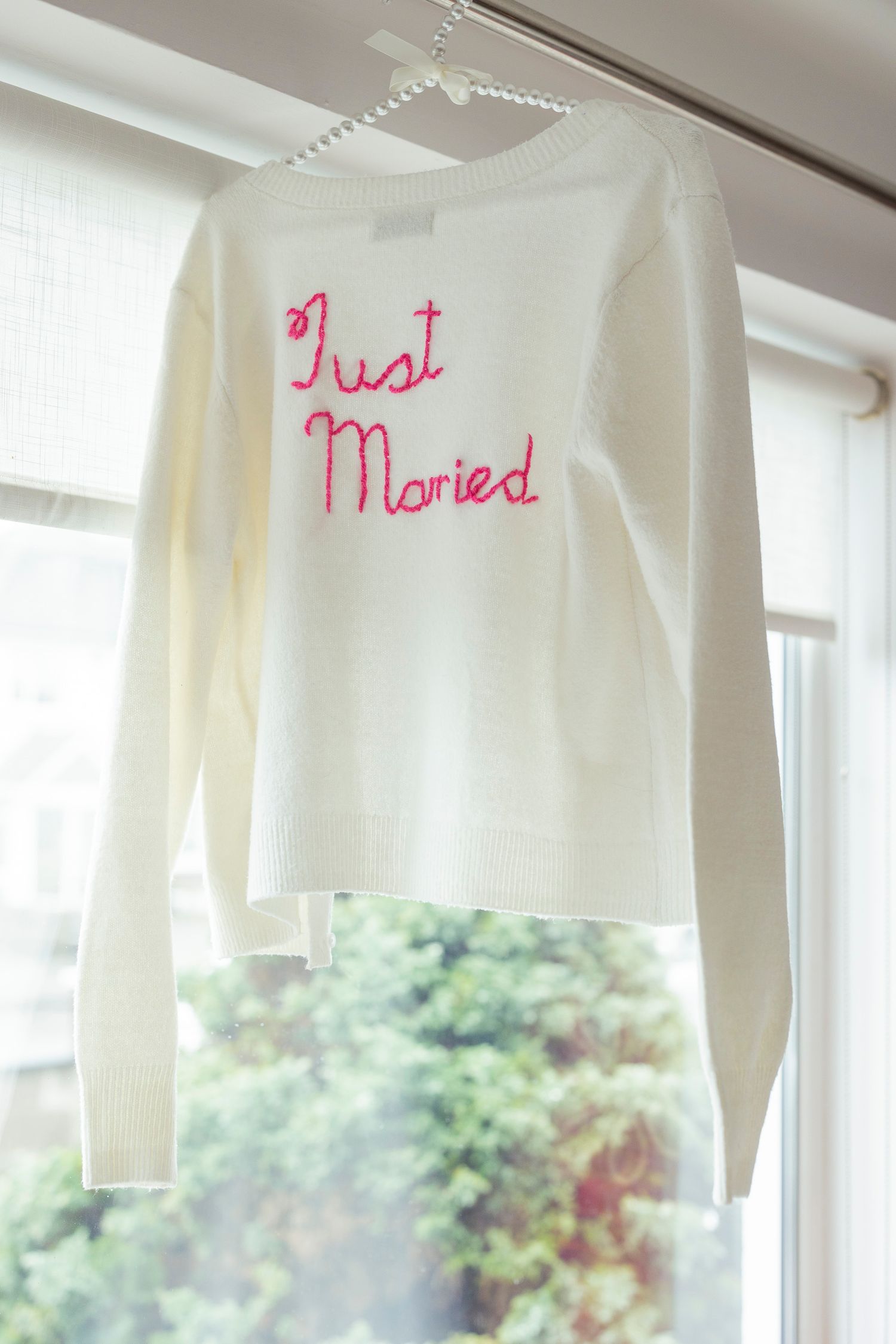 White knit sweater with pink embroidered Just Married text hanging by a window on pearl hanger.