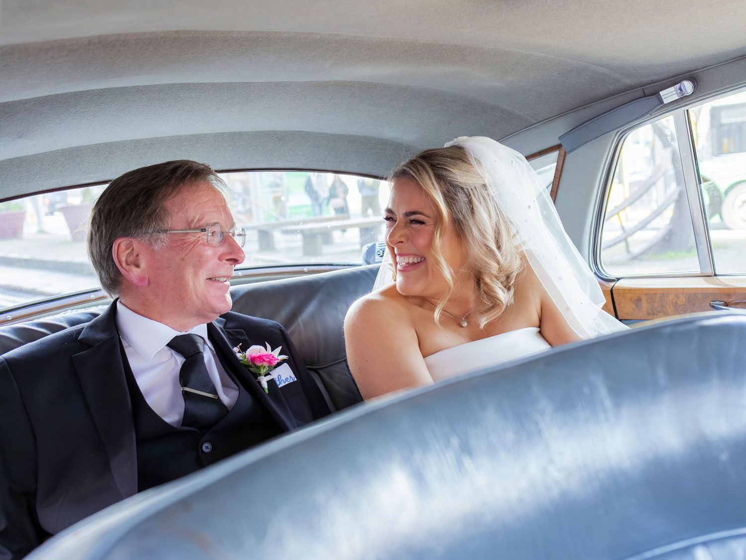 Smiling bride in veil and older gentleman in suit share a joyful moment in a classic vintage car.