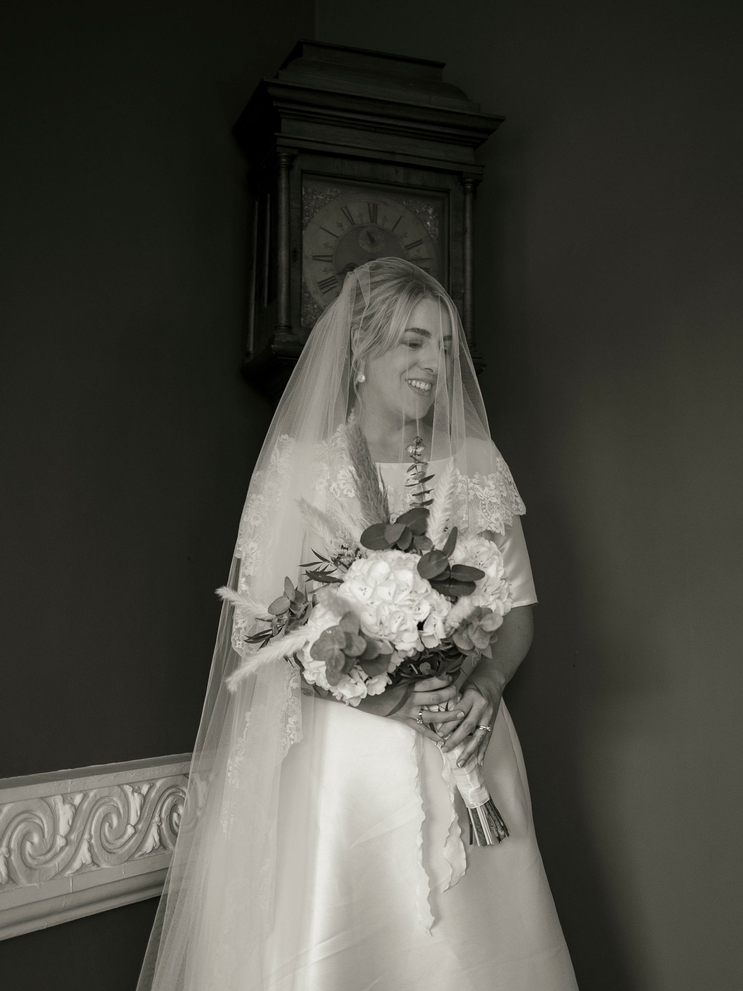 Black and white portrait of a smiling bride in veil holding a bouquet beside a grandfather clock.