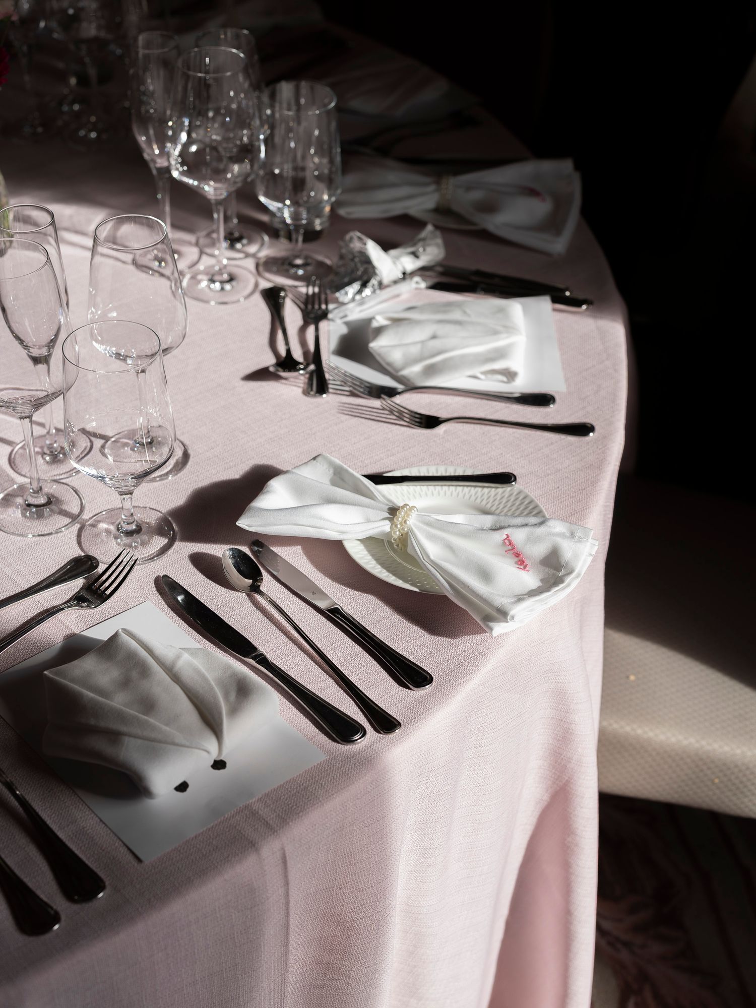 Elegant formal dining table set with white napkins, black cutlery, and crystal wine glasses on a pink tablecloth.