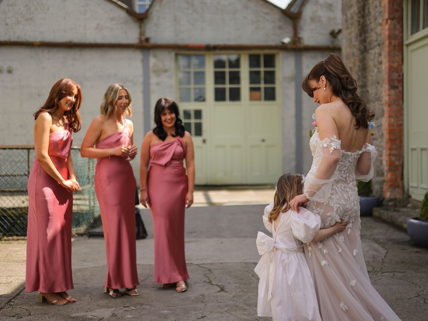 Bride in off-shoulder lace gown interacts with flower girl while bridesmaids in rose pink dresses look on.