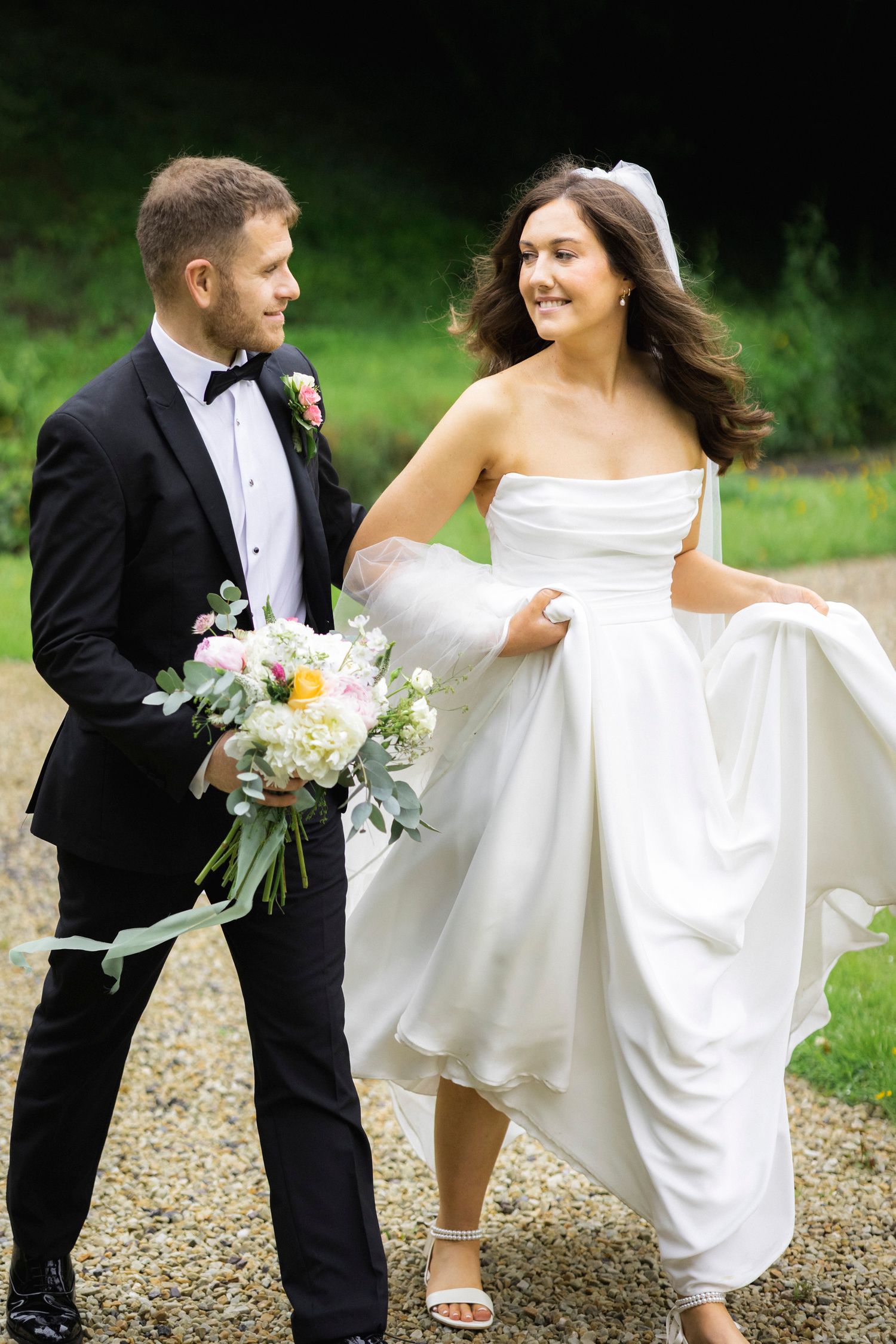 Smiling bride in white strapless gown walks with groom in black tuxedo holding floral bouquet on gravel path.