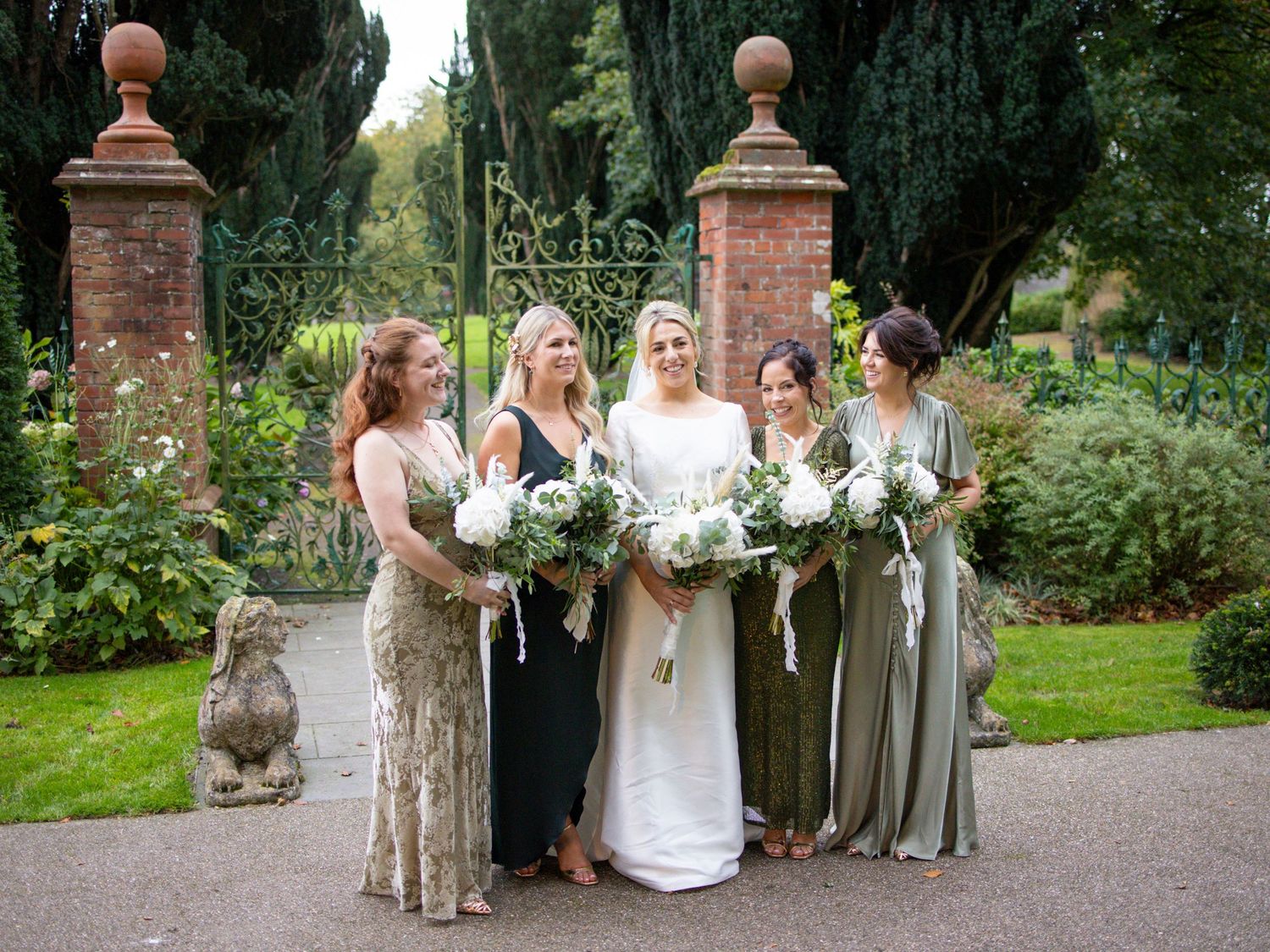 Bride with four bridesmaids in green and black dresses holding white floral bouquets at garden gate.