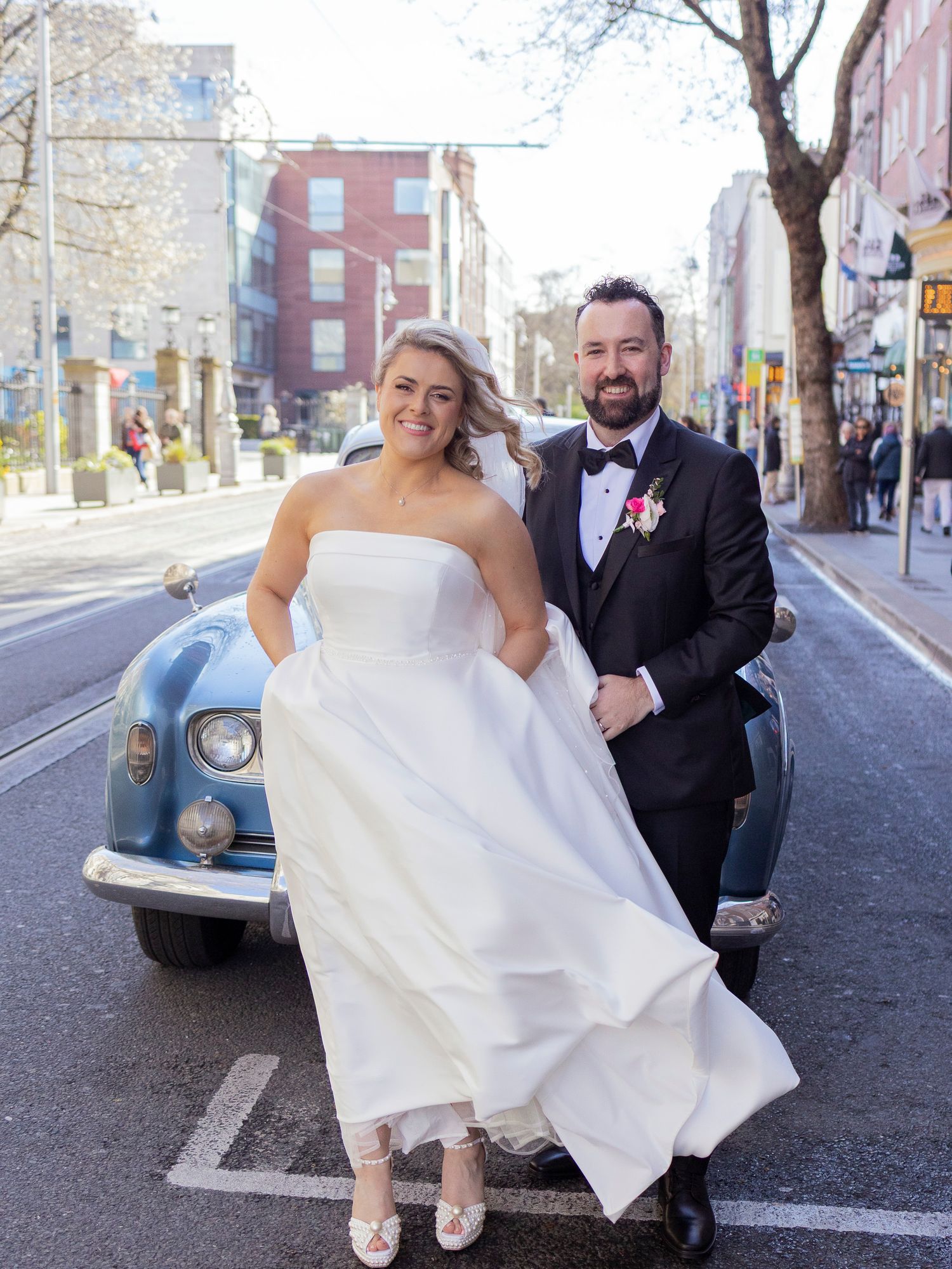 Bride and groom pose in front of vintage car on Dublin city street in wedding attire.