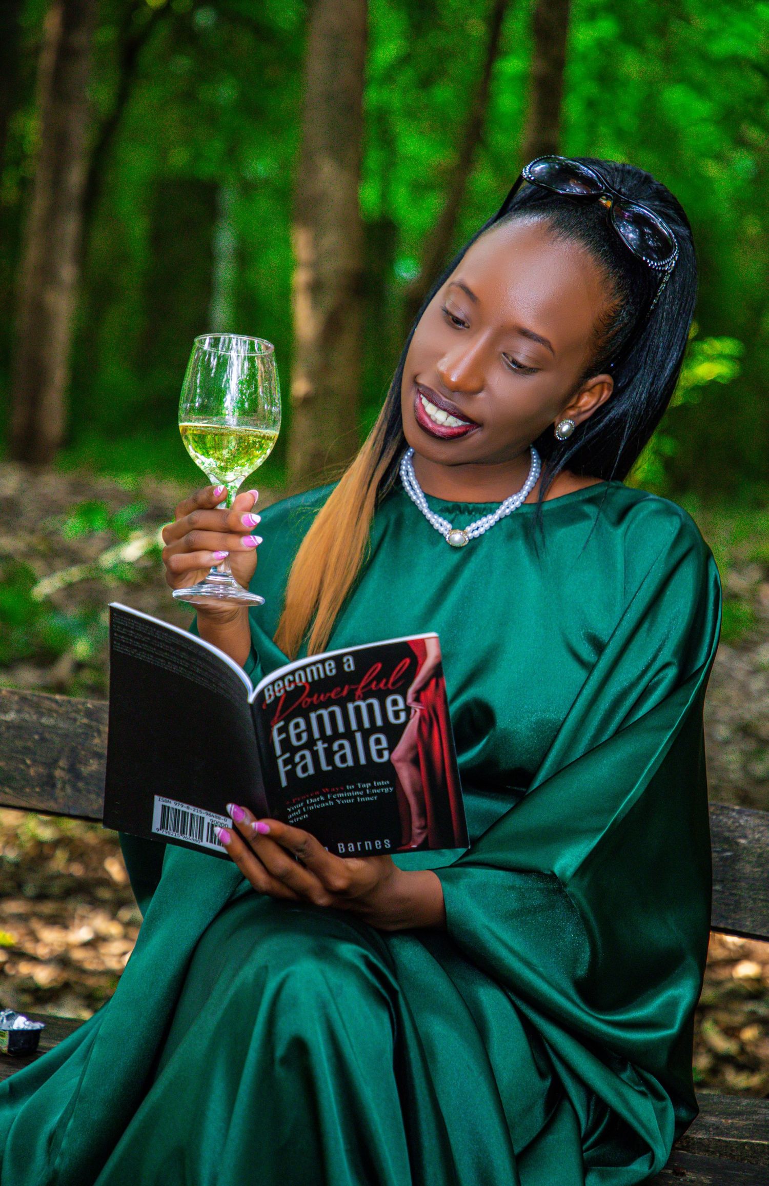 A woman in an elegant emerald green silk dress sitting on a park bench, holding a glass of white wine and reading a book titled Femme Fatal.