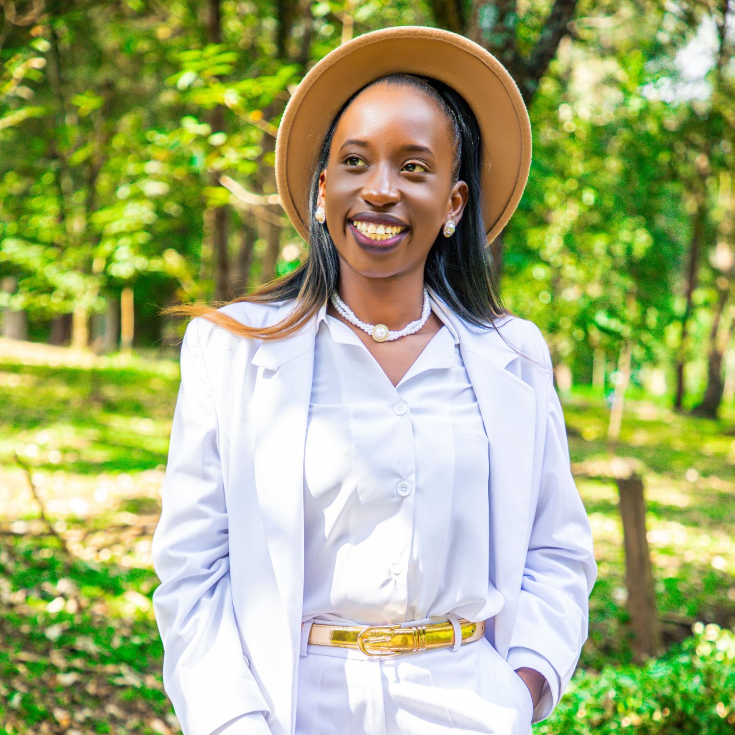 A smiling woman posing for an outdoor professional portrait in a white pantsuit, gold belt, and a beige fedora hat surrounded by lush green