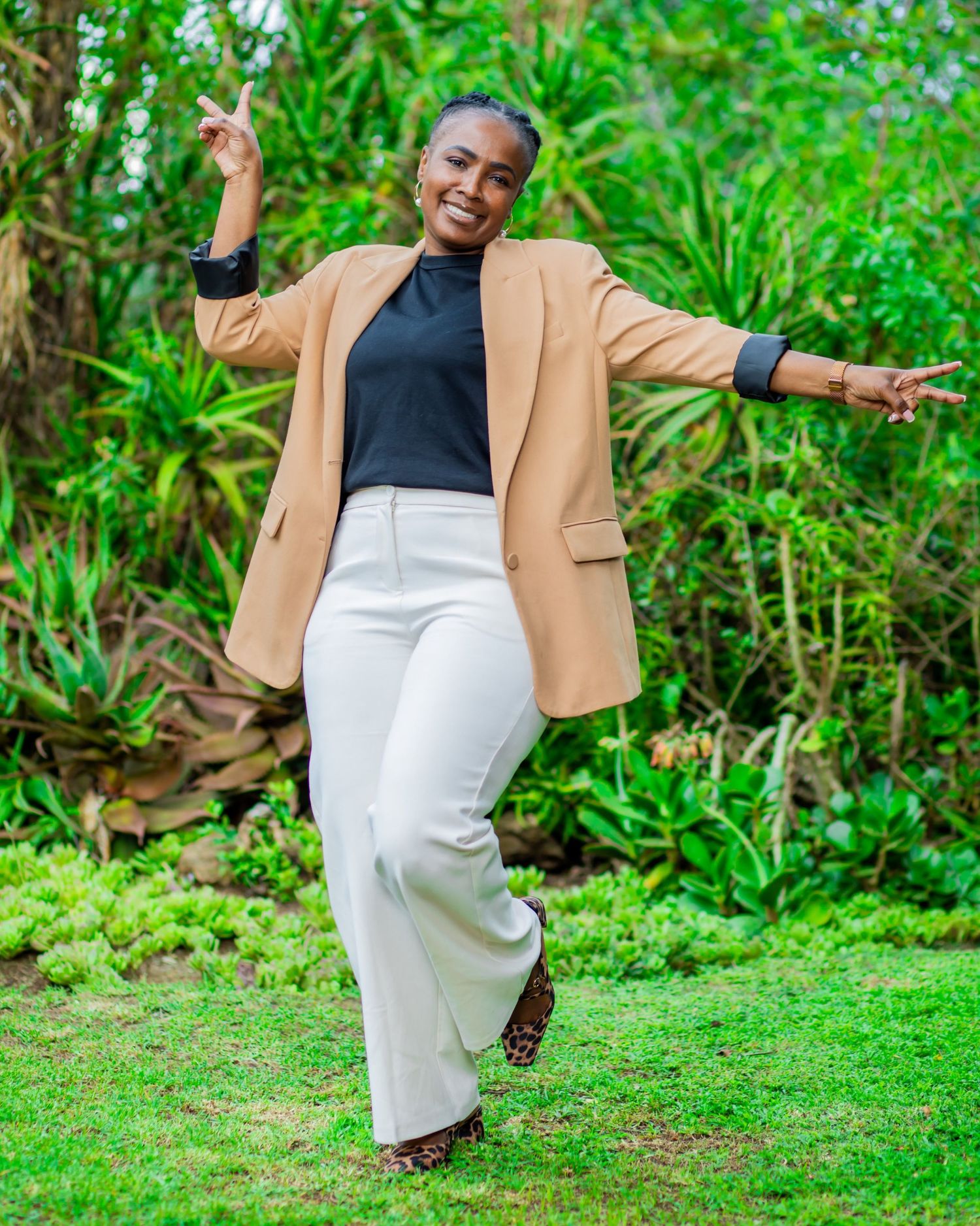 Full-length lifestyle portrait of a happy professional woman posing playfully, throwing peace signs on a green lawn.