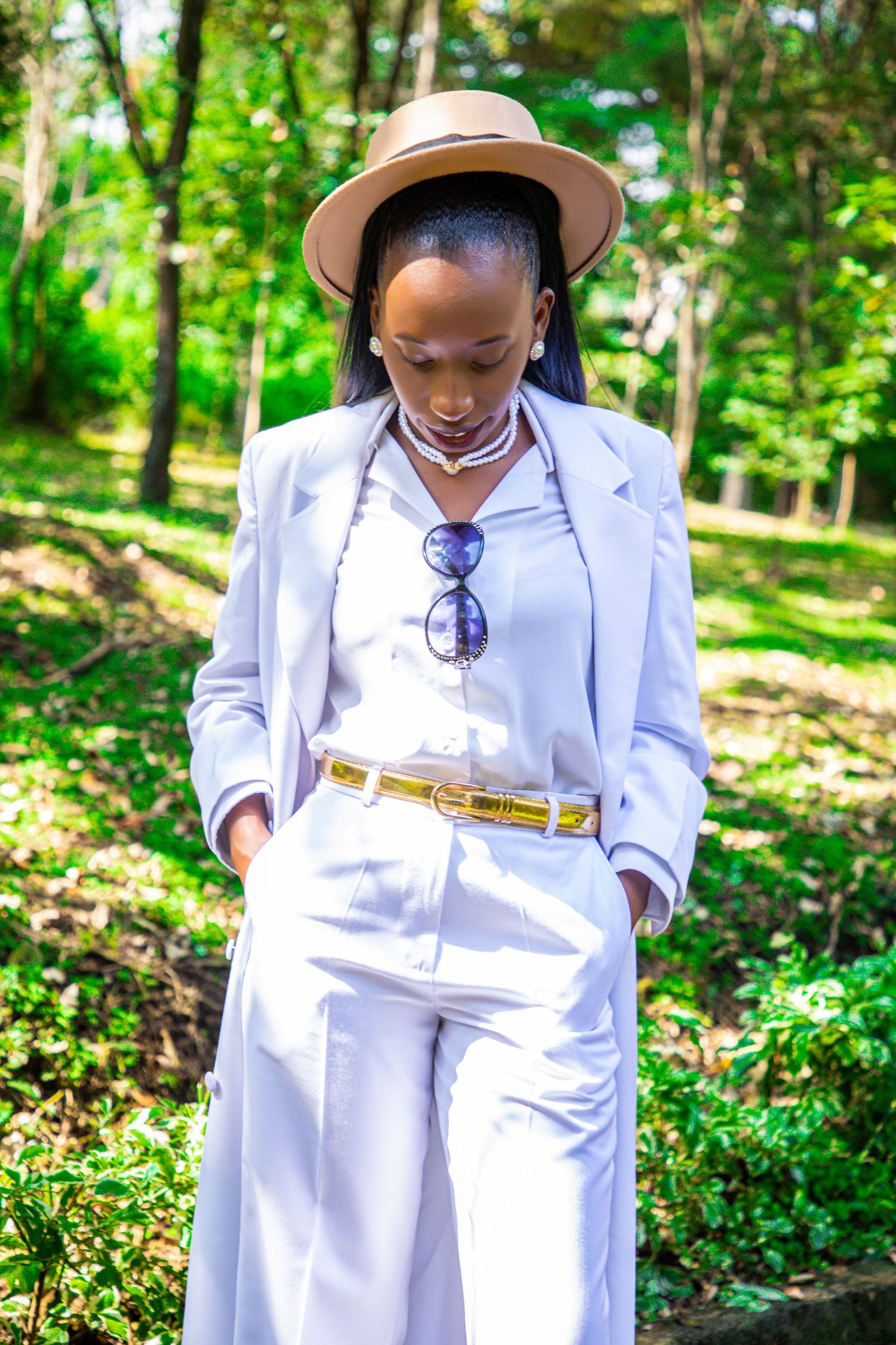 Full-length candid portrait of a woman in a white pantsuit and a brown fedora hat looking down with hands in pockets, posing outdoors.
