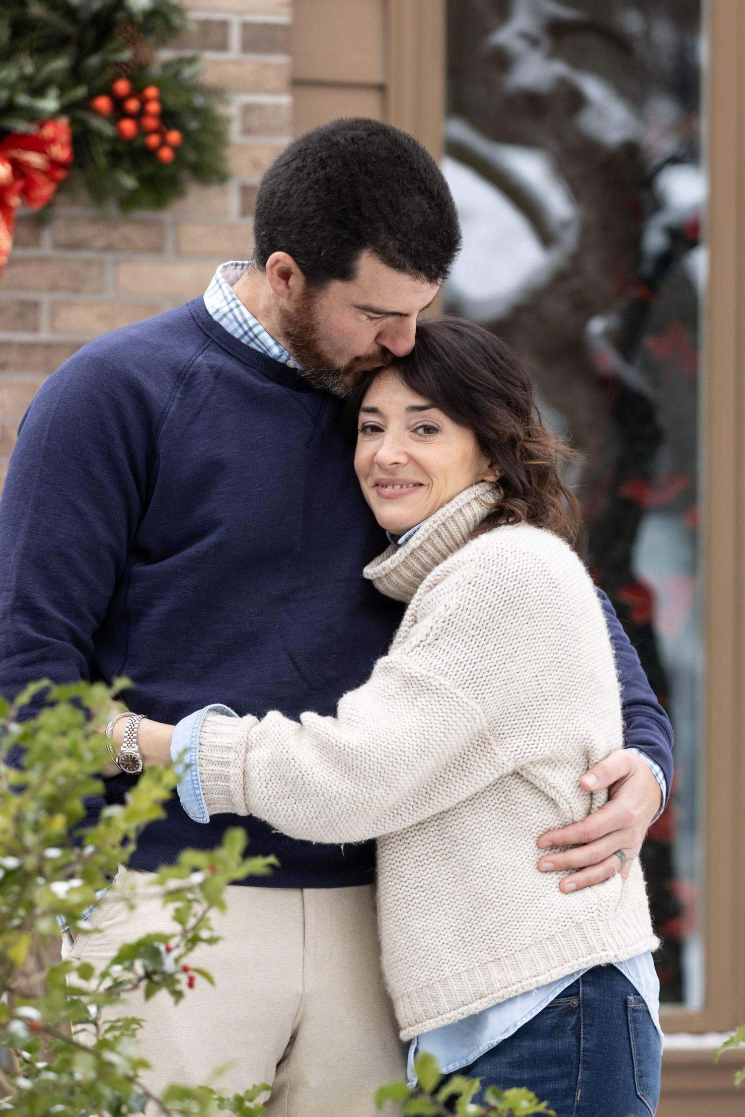 Couple in winter sweaters embracing warmly outside a holiday-decorated brick home.