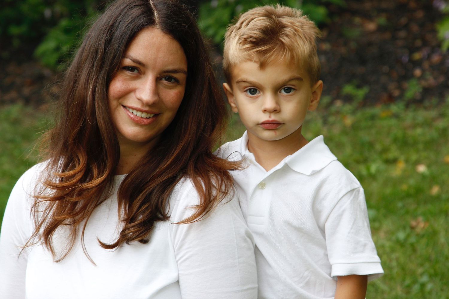 Mother and young son in white shirts posing together on a grassy outdoor background.