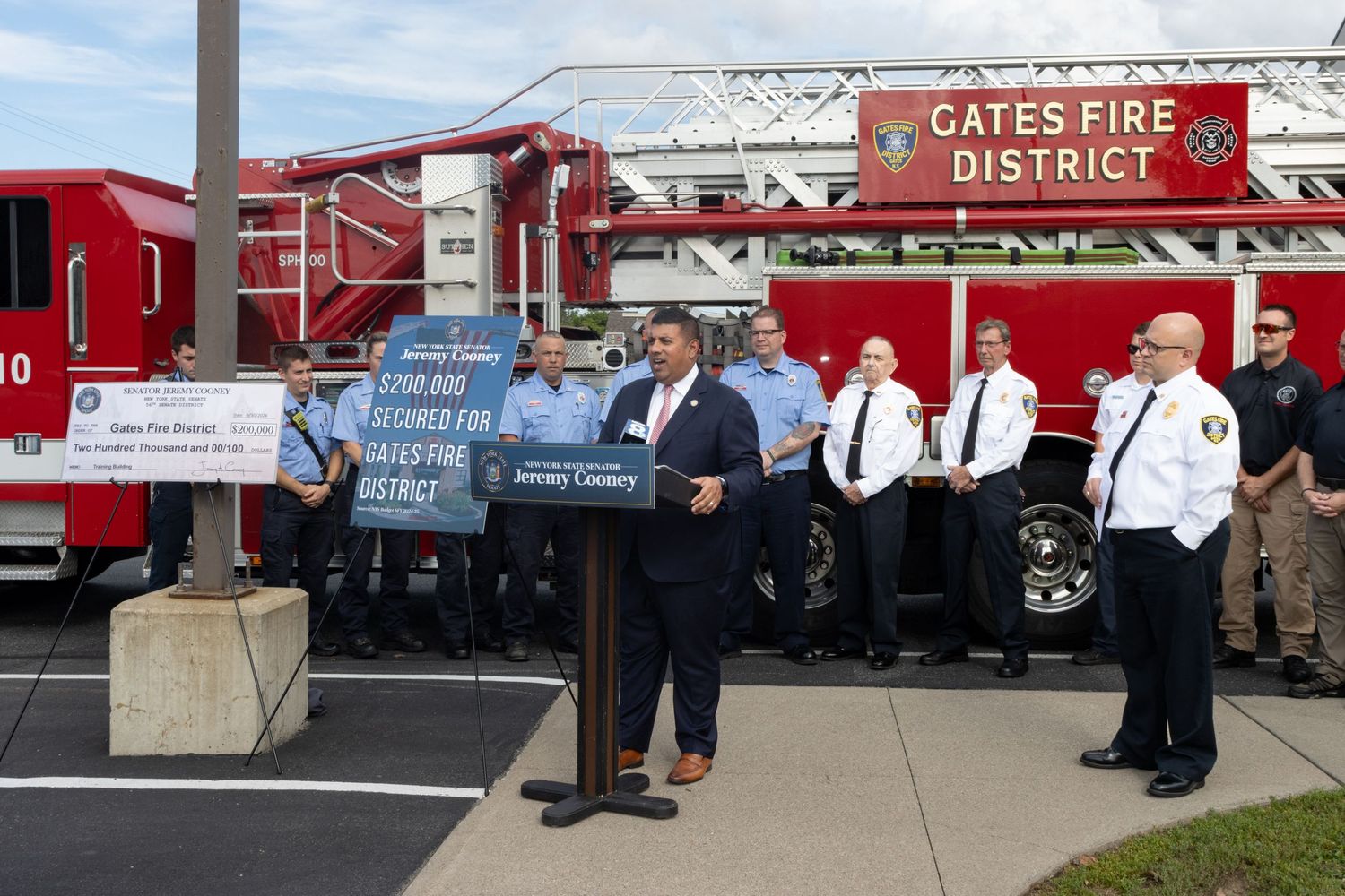 Official speaking at Gates Fire District podium with firefighters and fire truck behind him.