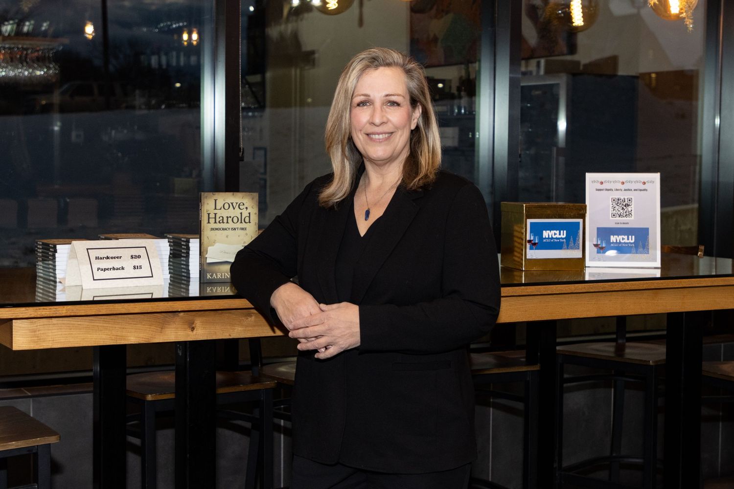 Woman in black blazer posing at a restaurant bar with wooden countertop and large windows.