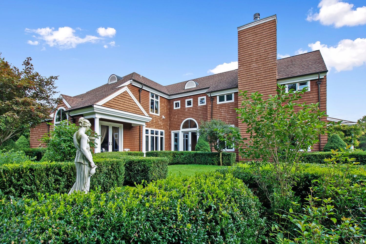 Elegant brick estate with manicured hedges, classical statue, arched windows, and sprawling landscaped gardens.