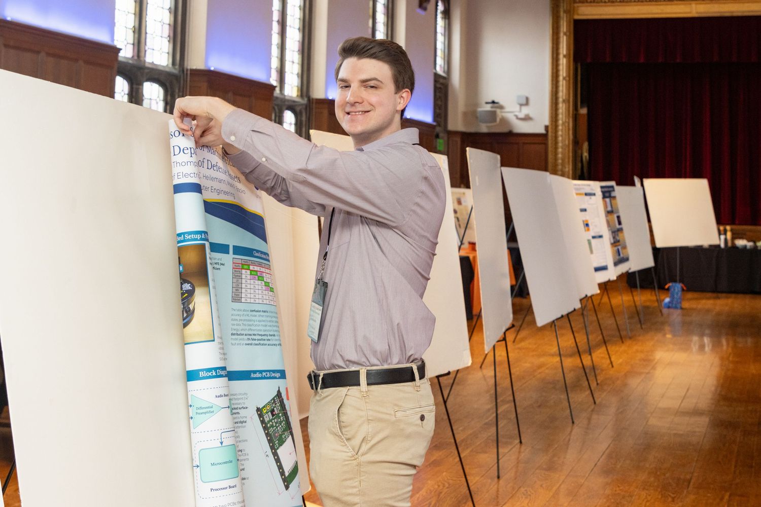 Young man setting up research poster display at an academic symposium in a historic hall.