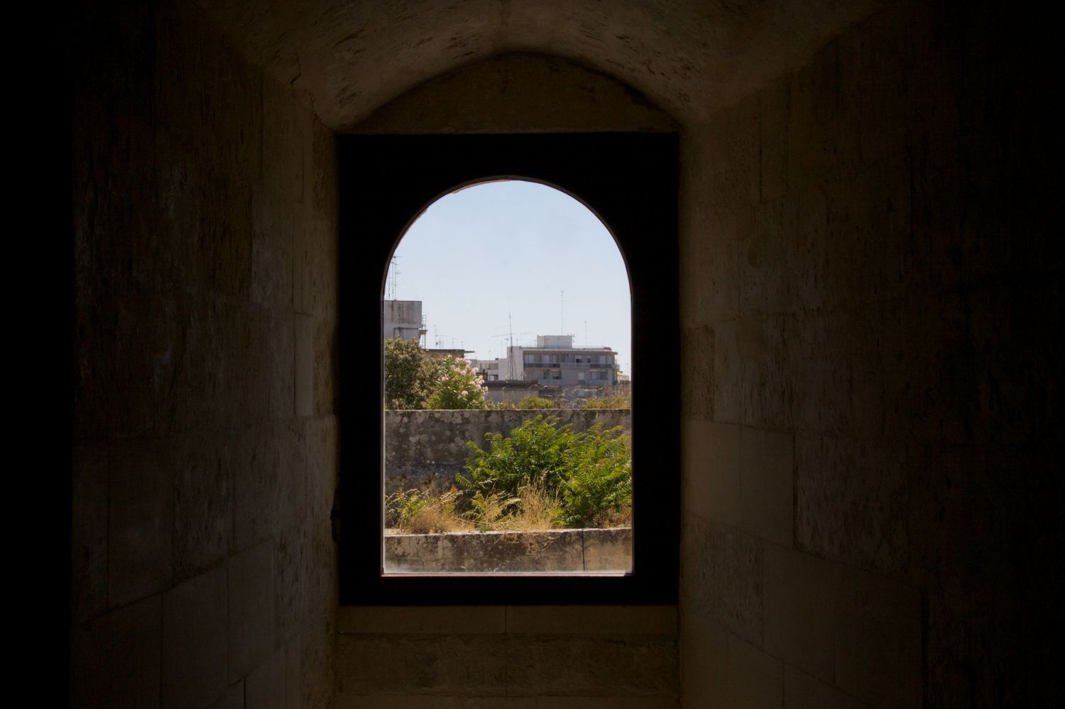 Arched stone window framing a sunny view of ancient walls and green trees outside.