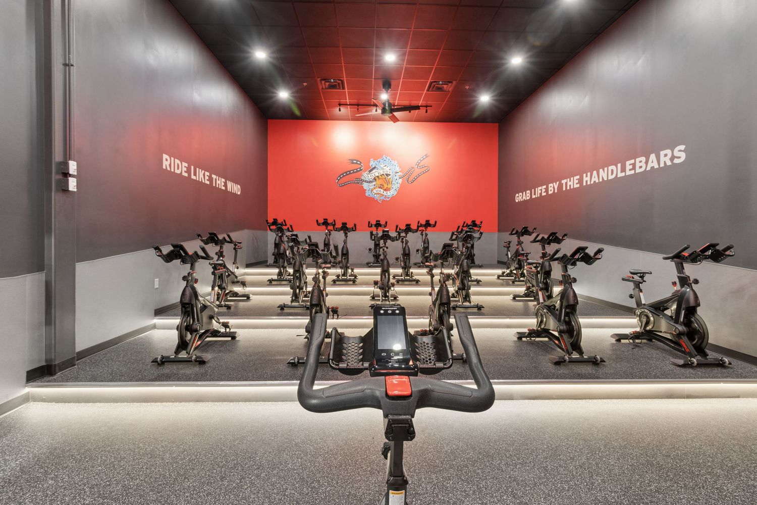 Indoor cycling studio with rows of stationary bikes, red accent wall, and motivational text in a modern fitness center.