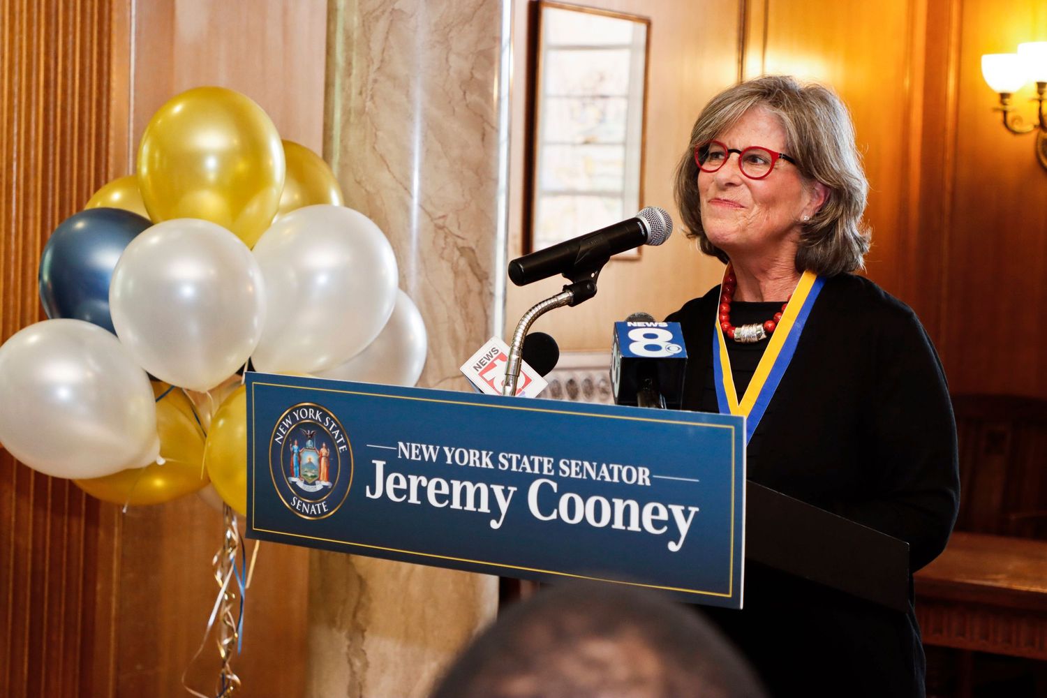 Woman speaking at a New York State Senator Jeremy Cooney podium with balloons in background.