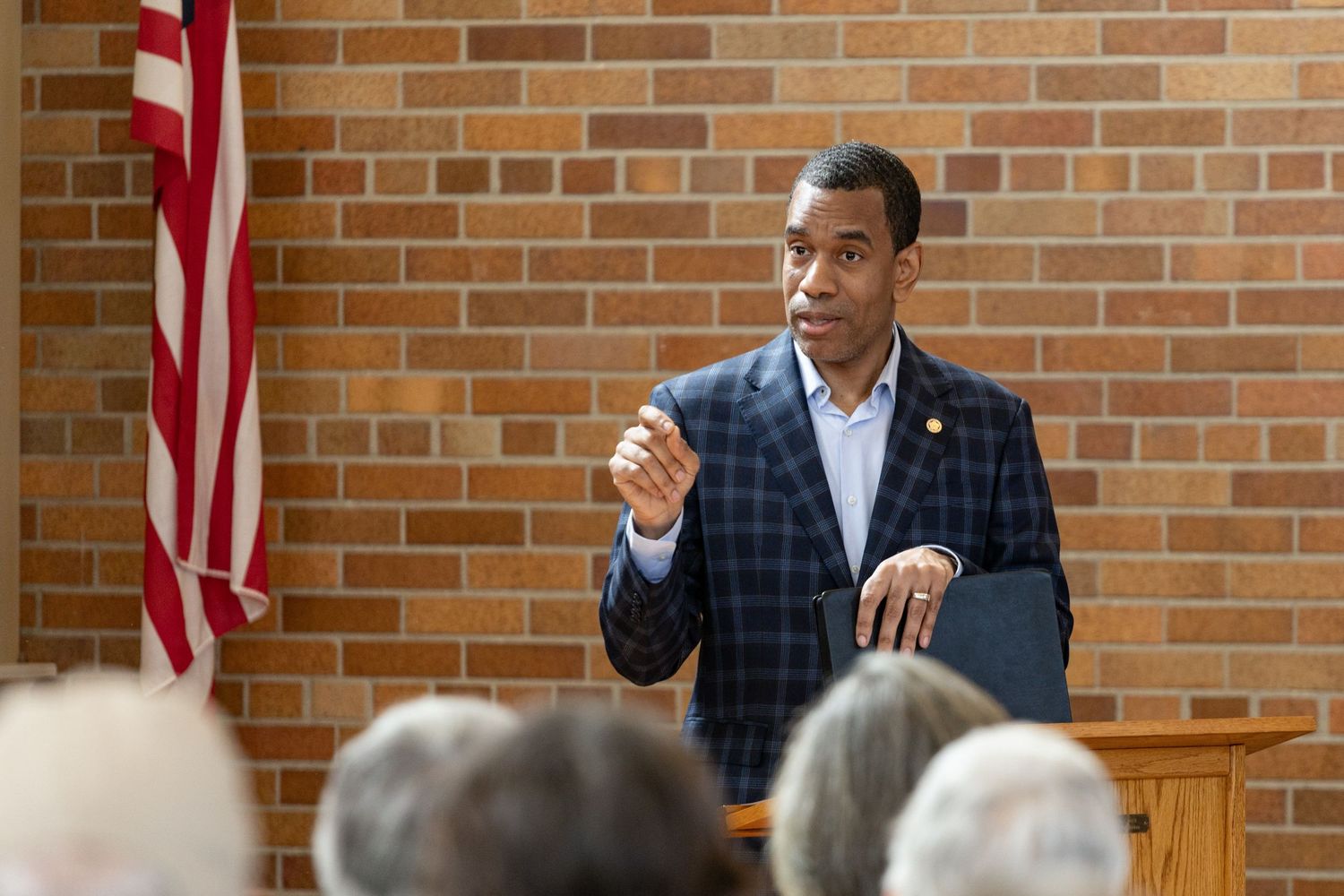 Man in plaid blazer speaking at podium before audience with American flag in background.
