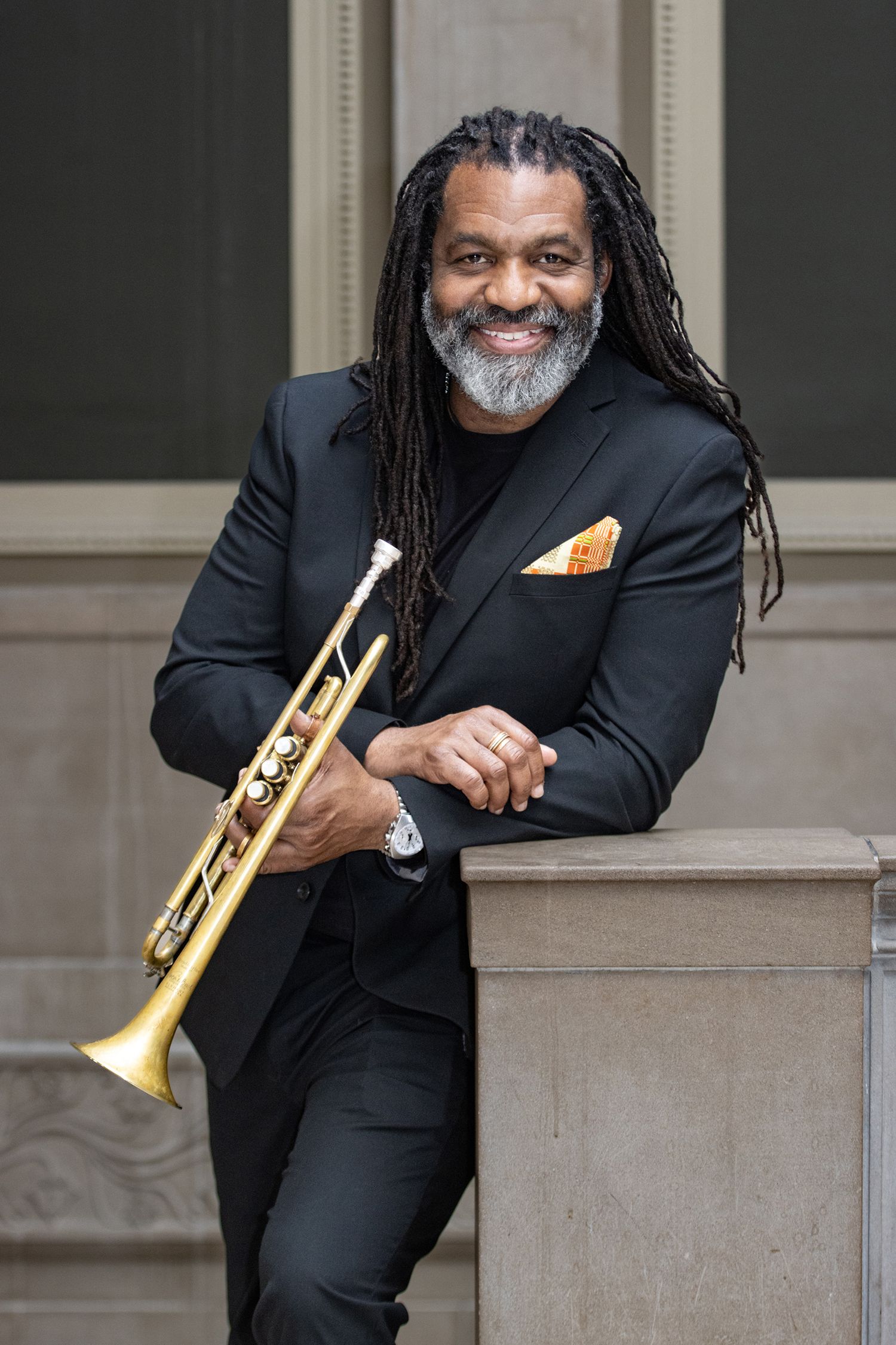 Smiling musician in black suit holding trumpet while leaning against stone pillar outdoors.