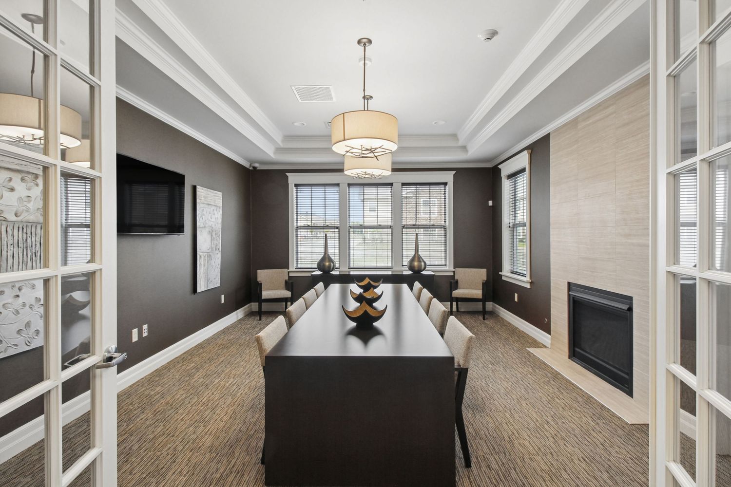 Elegant conference room with dark wood table, tray ceiling, modern chandelier, fireplace, and large windows in neutral tones.