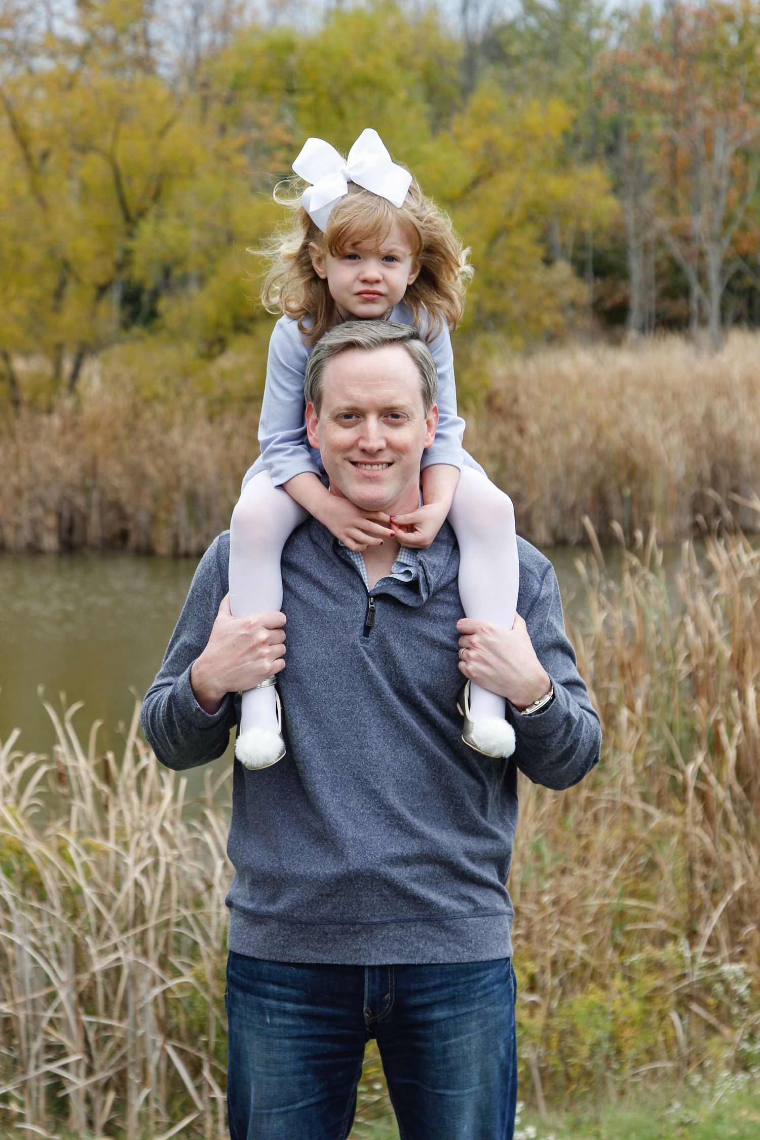 Smiling father carrying young daughter with white bow on his shoulders near a pond.
